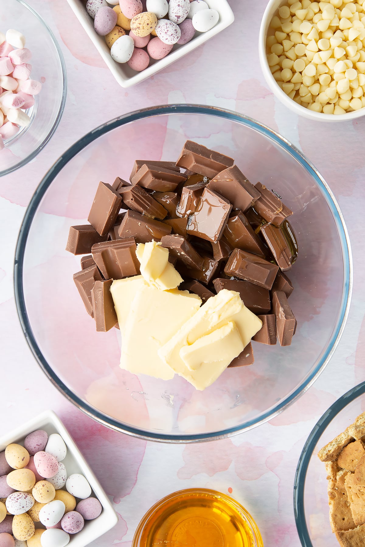 Milk chocolate, butter and golden syrup in a glass mixing bowl. Ingredients to make Mini Egg rocky road surrounds the bowl.