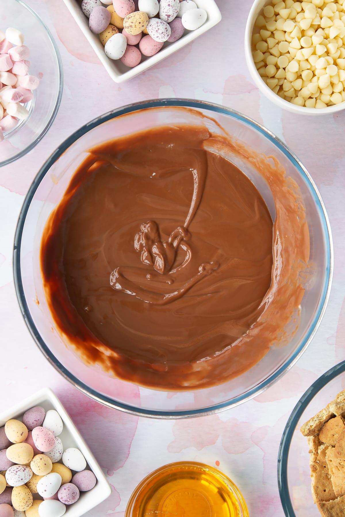 Melted milk chocolate, butter and golden syrup in a glass mixing bowl. Ingredients to make Mini Egg rocky road surrounds the bowl.