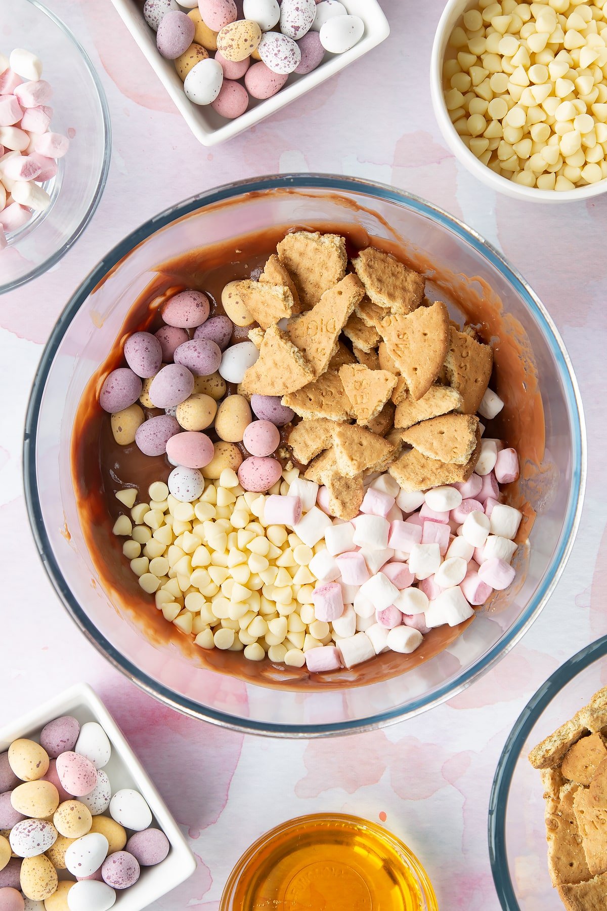 Melted milk chocolate, butter and golden syrup in a glass mixing bowl topped with white chocolate chips, digestives, marshmallows and Mini Eggs. Ingredients to make Mini Egg rocky road surrounds the bowl.