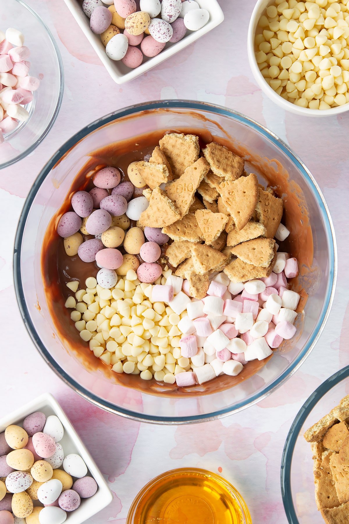 Melted milk chocolate, butter and golden syrup in a glass mixing bowl topped with white chocolate chips, digestives, marshmallows and Mini Eggs. Ingredients to make Mini Egg rocky road surrounds the bowl.