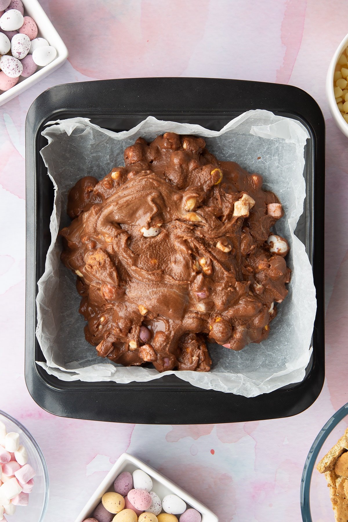 Rocky road mix in a tray lined with baking paper. Ingredients to make Mini Egg rocky road surrounds the tray.