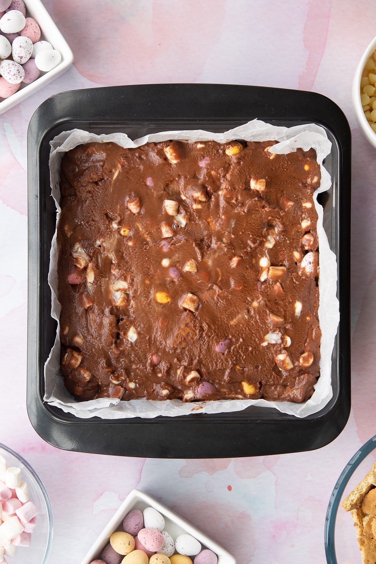 Rocky road mix levelled off in a tray lined with baking paper. Ingredients to make Mini Egg rocky road surrounds the tray.