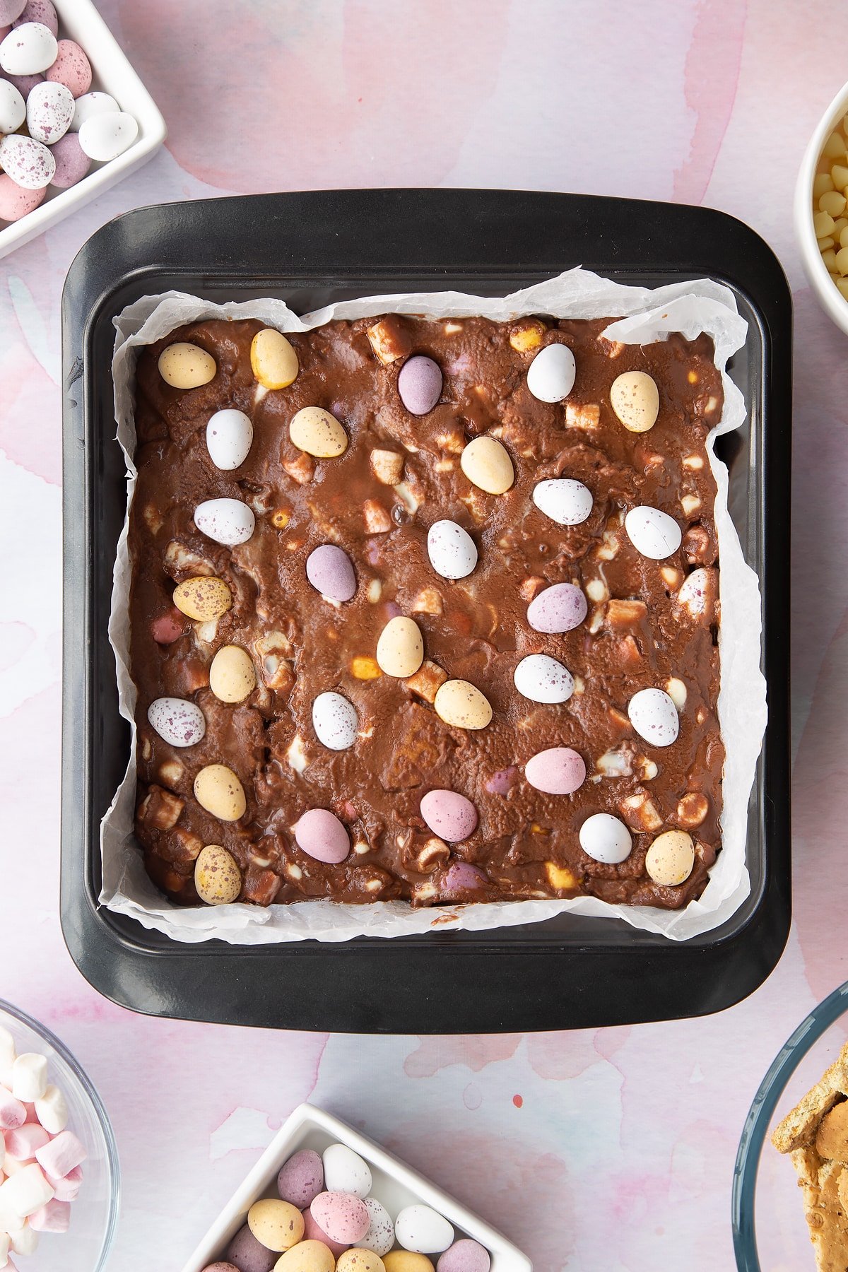 Rocky road mix levelled off in a tray and topped with Mini Eggs. Ingredients to make Mini Egg rocky road surrounds the tray.