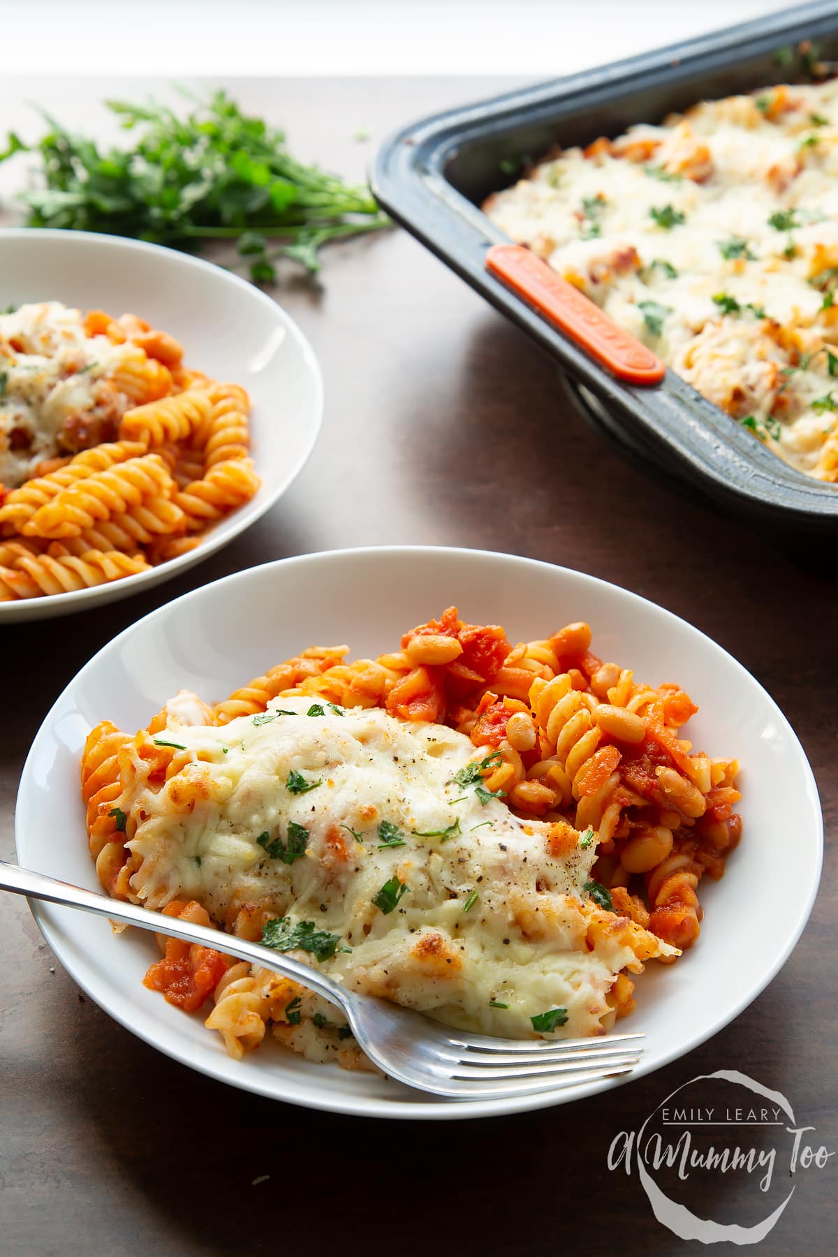 Pasta and baked beans served to white bowls with melted mozzarella. The tray of pasta bake is shown in the background