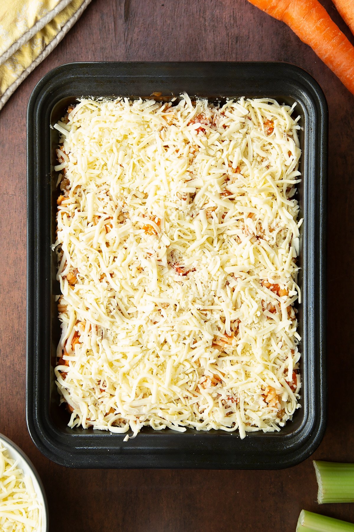 Baked beans and pasta in a tray, topped with shredded mozzarella. Ingredients to make pasta and baked beans surround the tray.