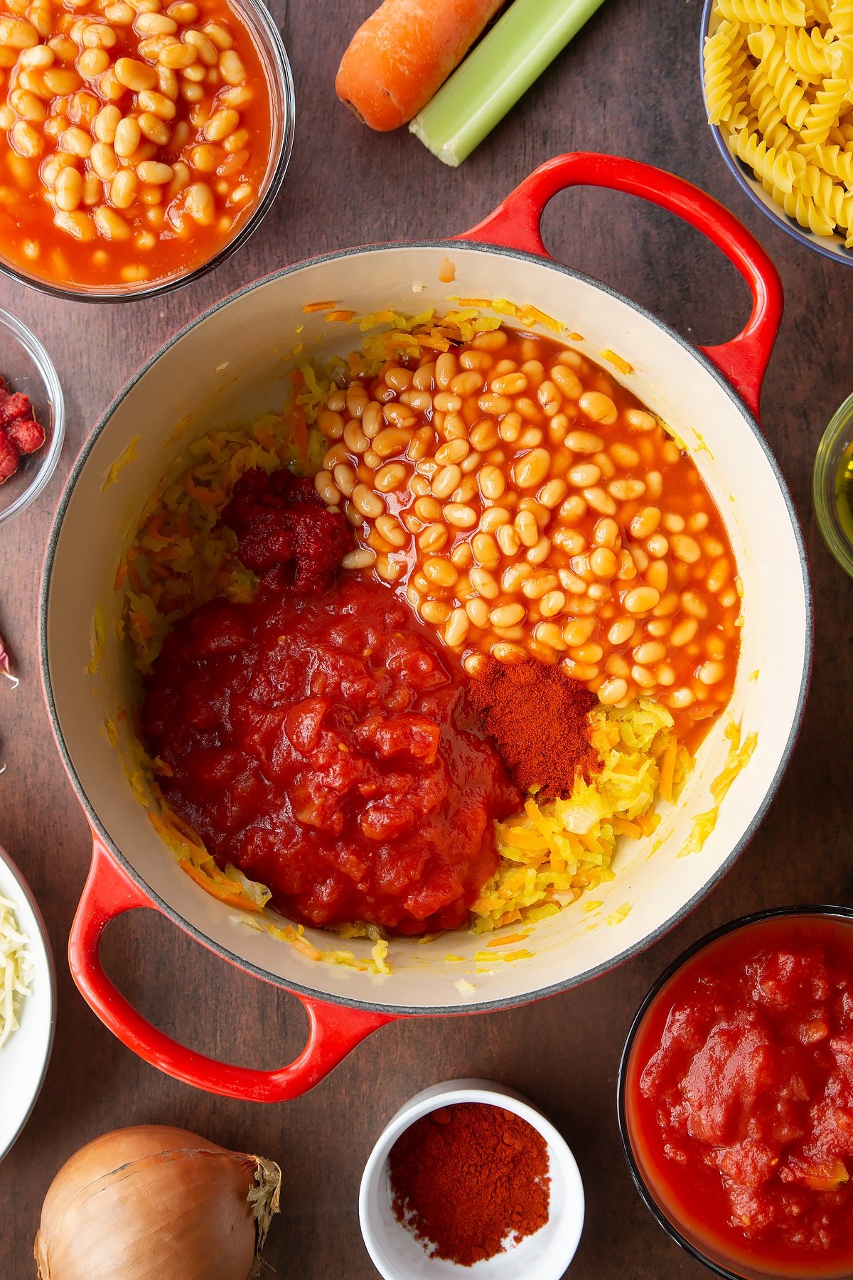 Fried grated veg in a saucepan with beans, tomatoes, tomato puree, paprika on top. Ingredients to make pasta and baked beans surround the pan.