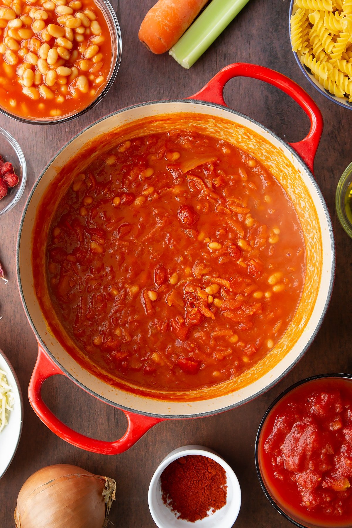 Grated veg, baked beans, tomatoes, tomato puree and paprika simmered down in a saucepan. Ingredients to make pasta and baked beans surround the pan.