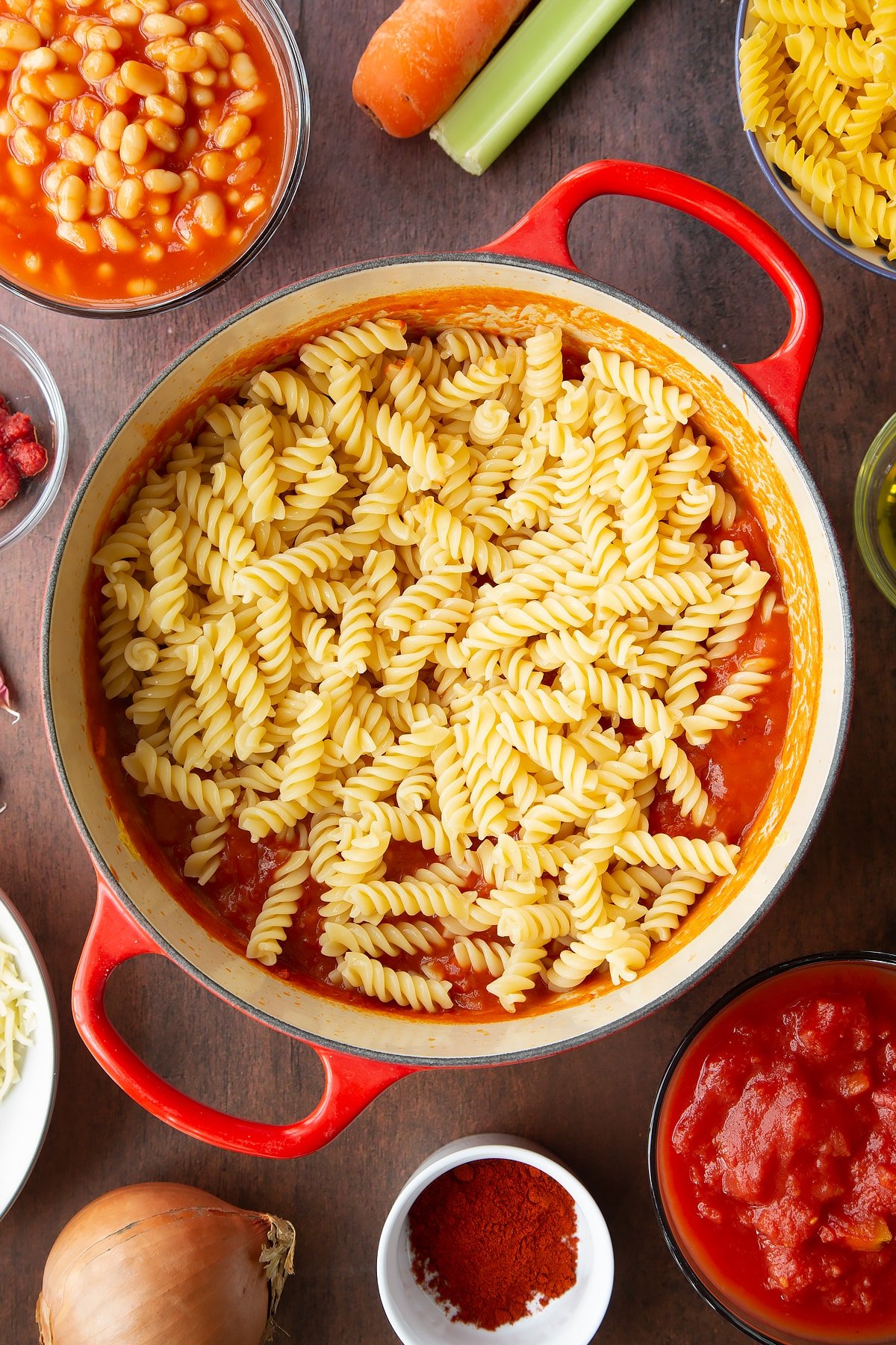 Grated veg, baked beans, tomatoes, tomato puree and paprika simmered down in a saucepan with pasta on top. Ingredients to make pasta and baked beans surround the pan.
