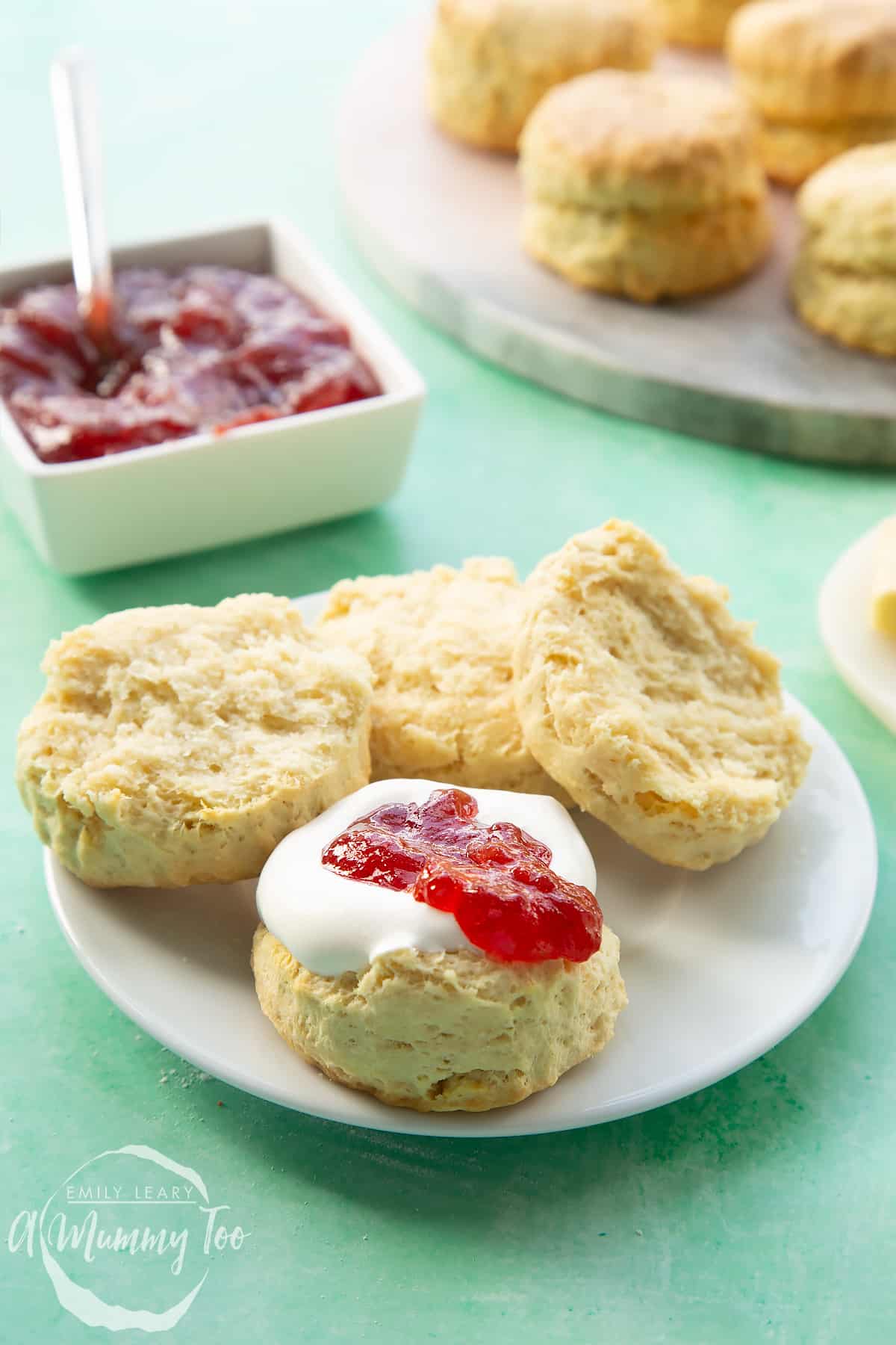 Two dairy free scones on a white plate. They are split in half and one is topped with jam and vegan whipped cream.