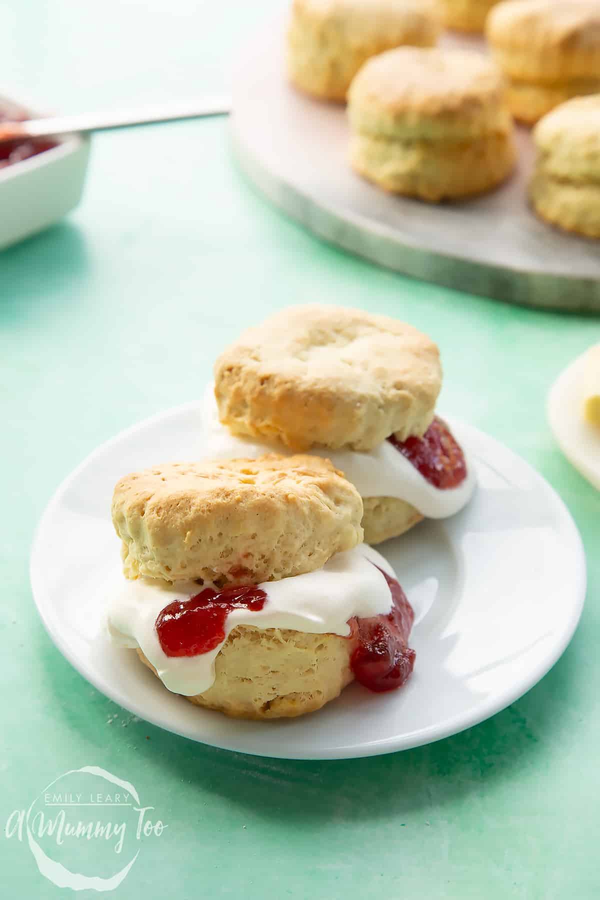 Two dairy free scones on a white plate. They are filled with jam and vegan whipped cream.