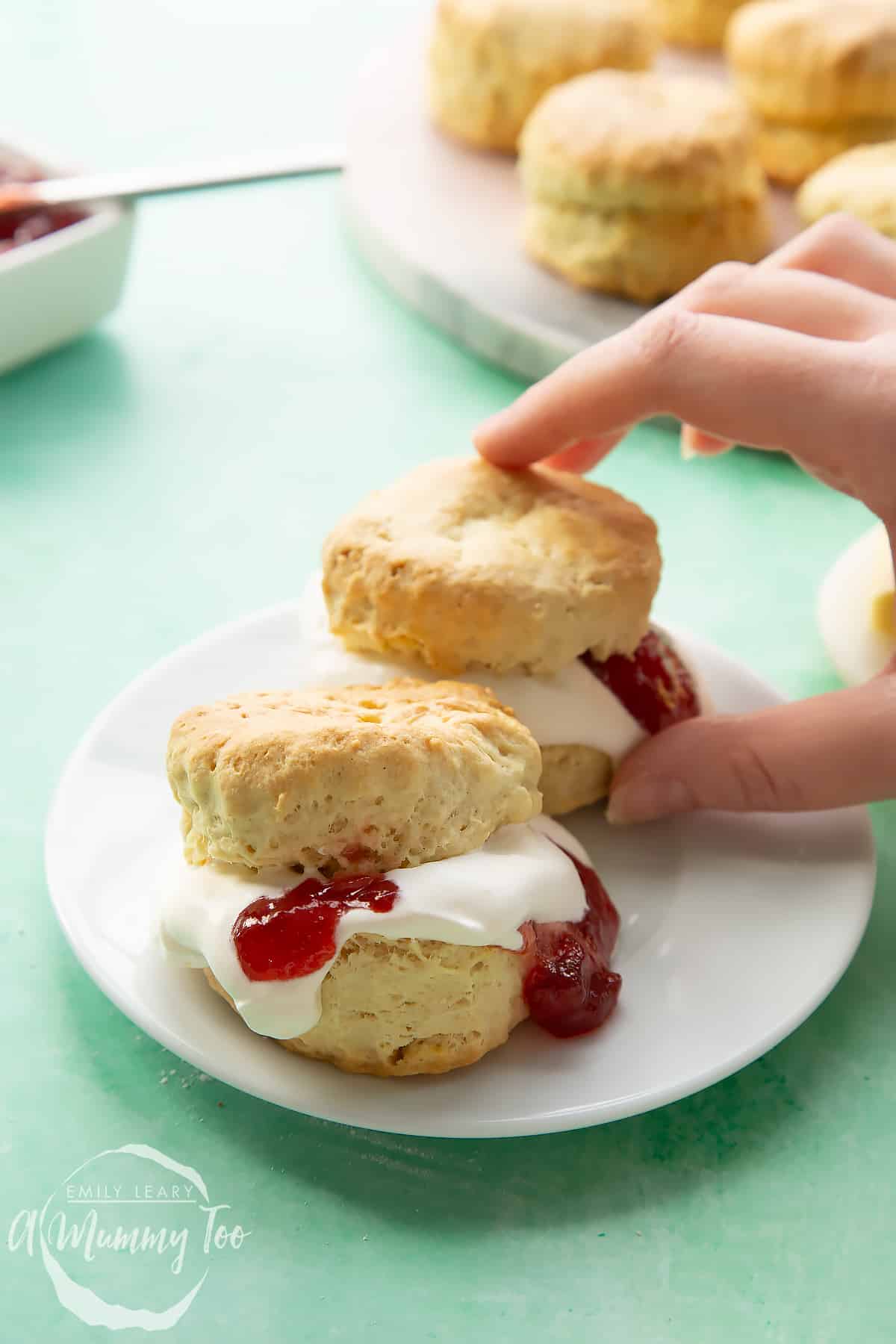 Two dairy free scones on a white plate. They are filled with jam and vegan whipped cream. A hand reaches for one.