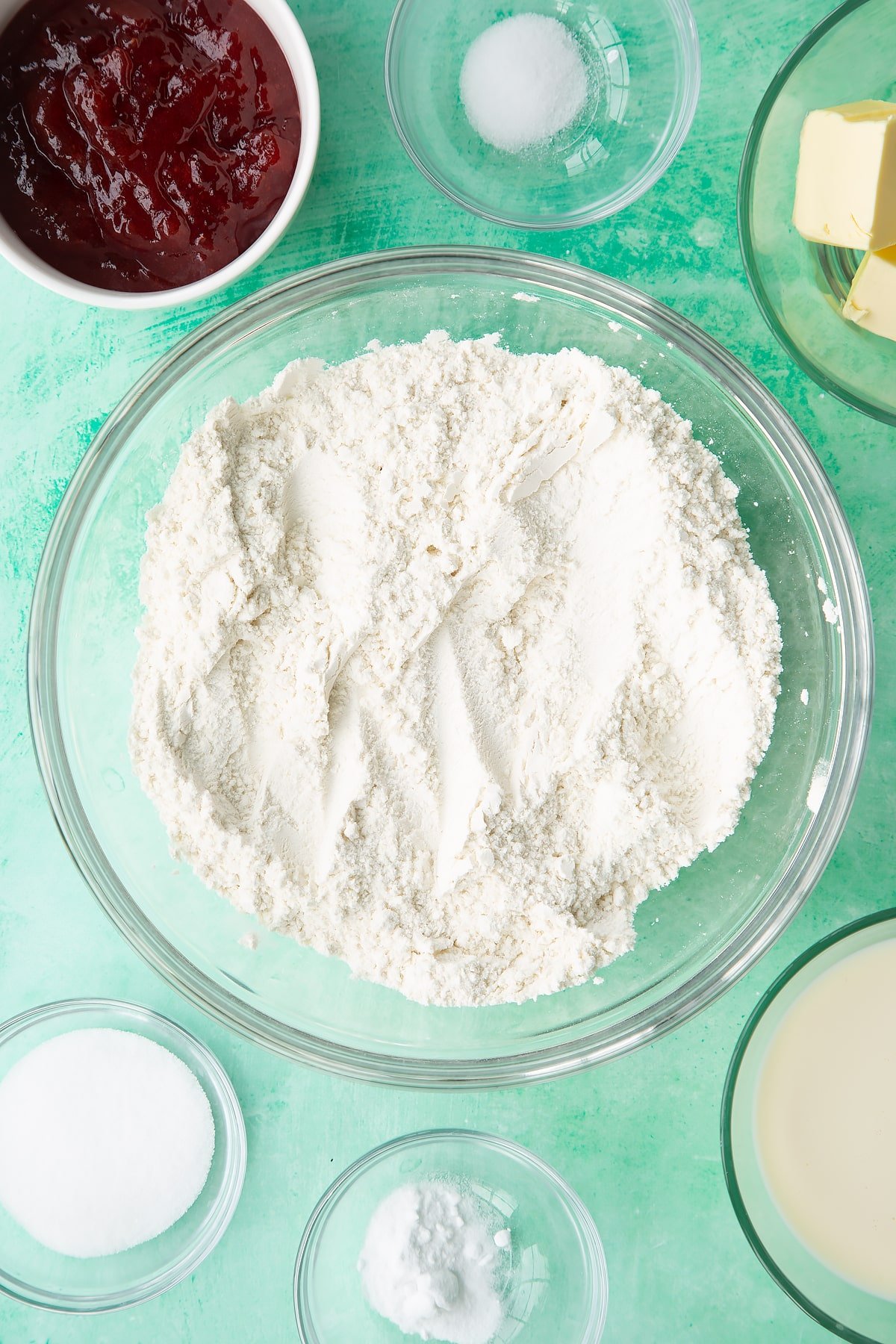 Flour, sugar, salt and bicarbonate of soda mixed together in a bowl. Ingredients to make dairy free scones surround the bowl.