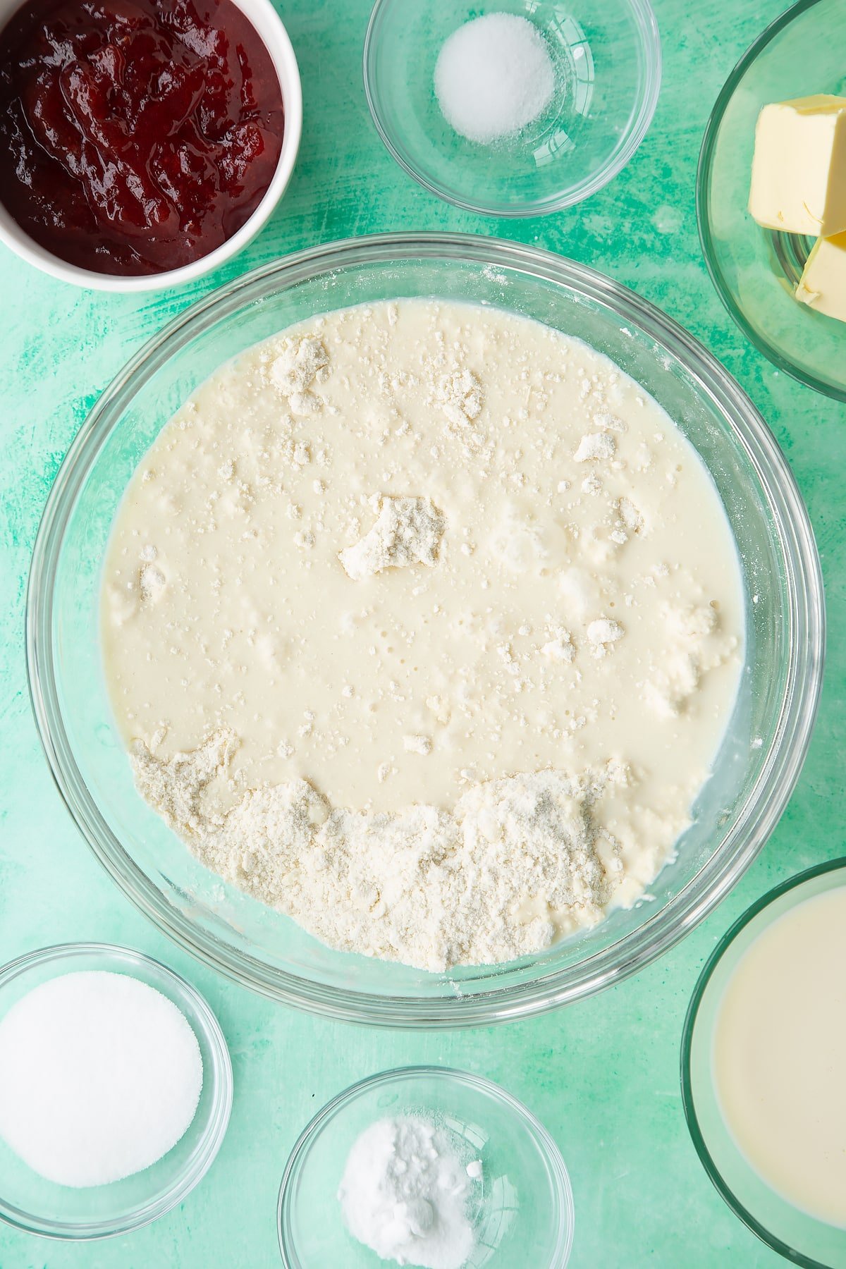 Flour, sugar, salt, bicarbonate of soda and vegan butter rubbed together in a bowl with plant milk on top. Ingredients to make dairy free scones surround the bowl.