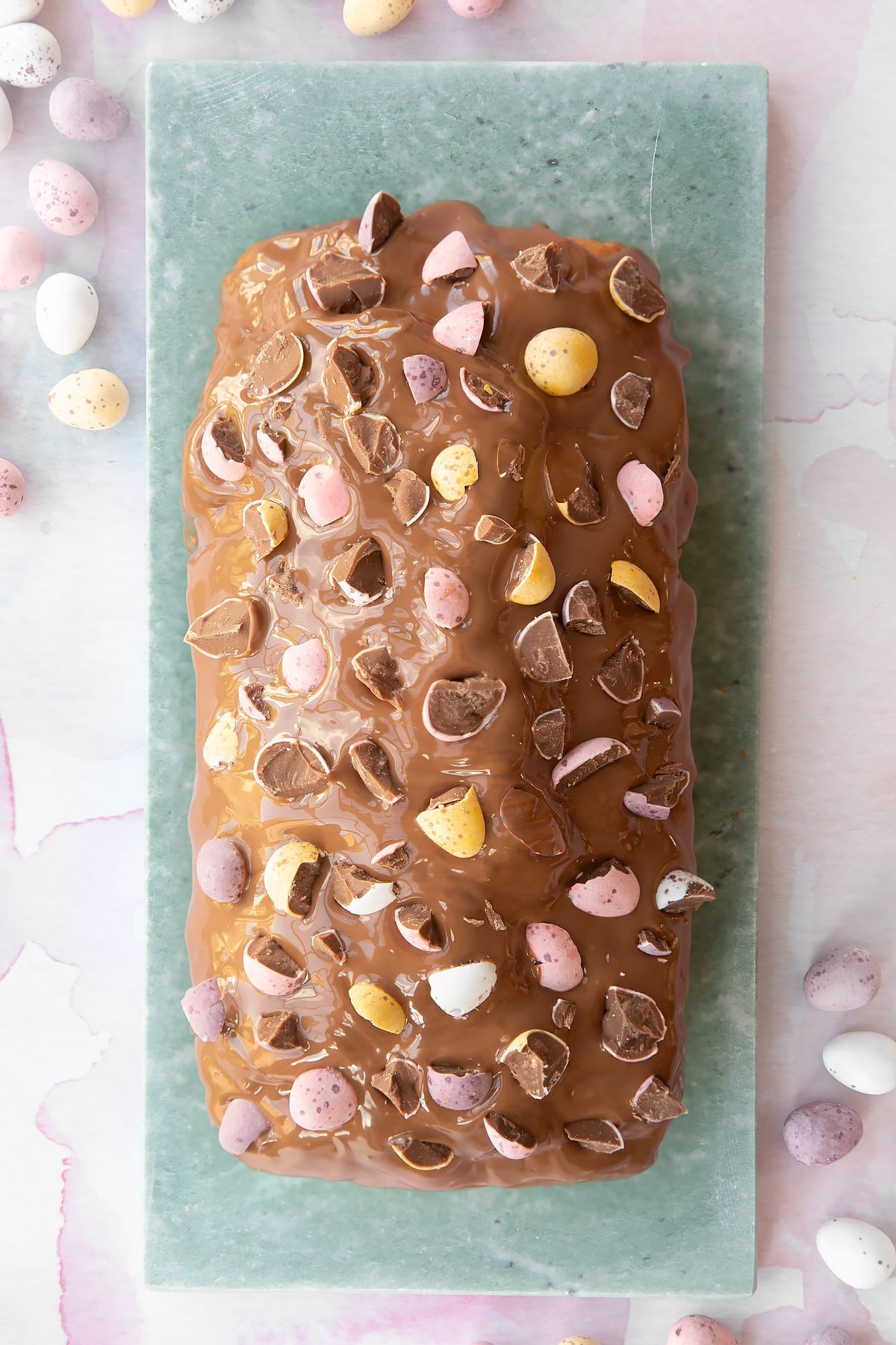 Easter banana bread decorated with milk chocolate and chopped Mini Eggs on a long grey marble board.