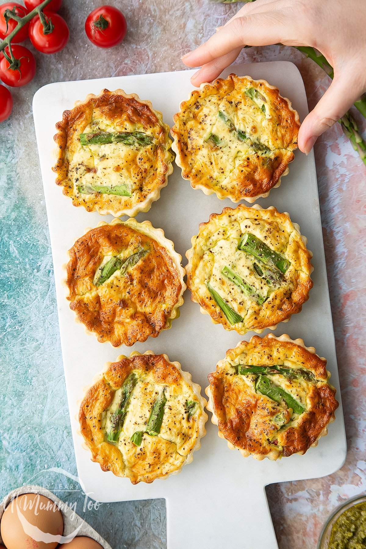 Six asparagus tartlets on a white marble board, shown from above. A hand reaches for one.