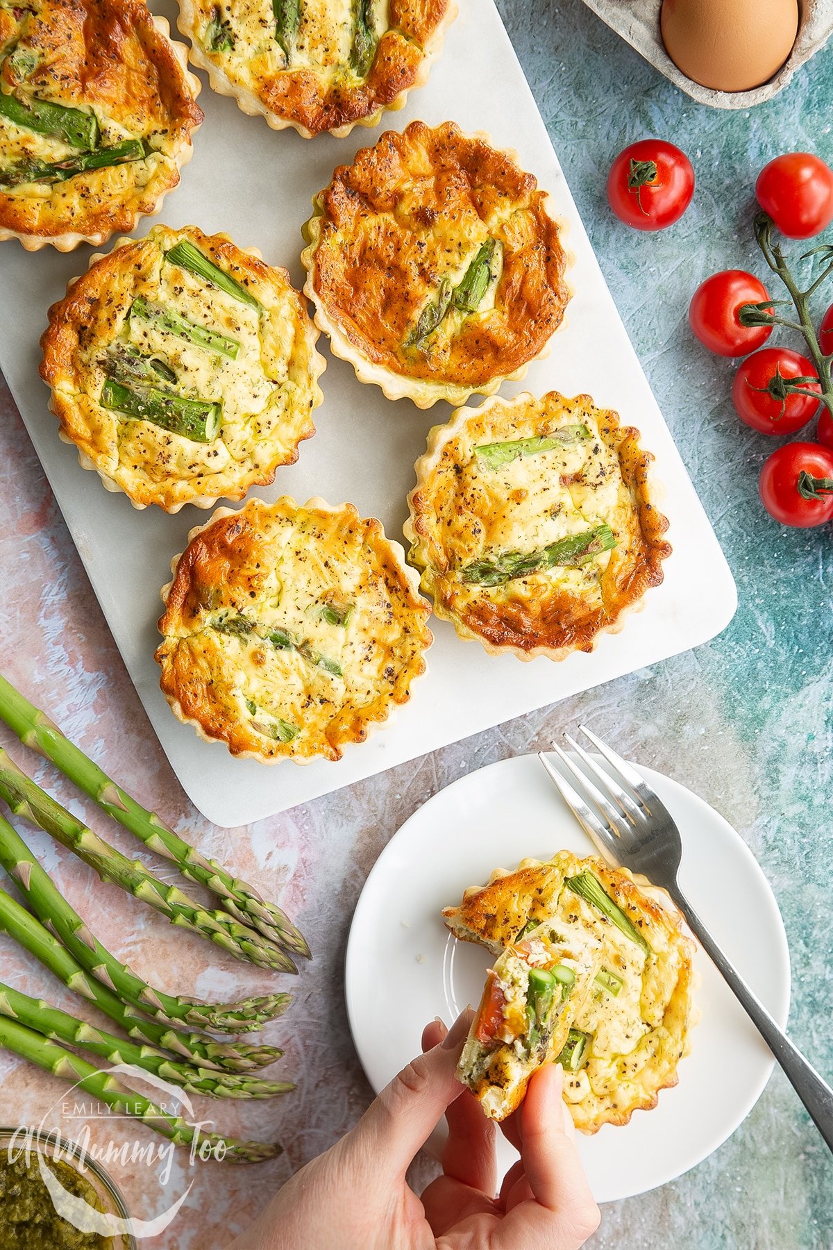 Asparagus tartlets on a white marble board. One is on a white plate with a fork and a hand holds a piece. Shown from above. 