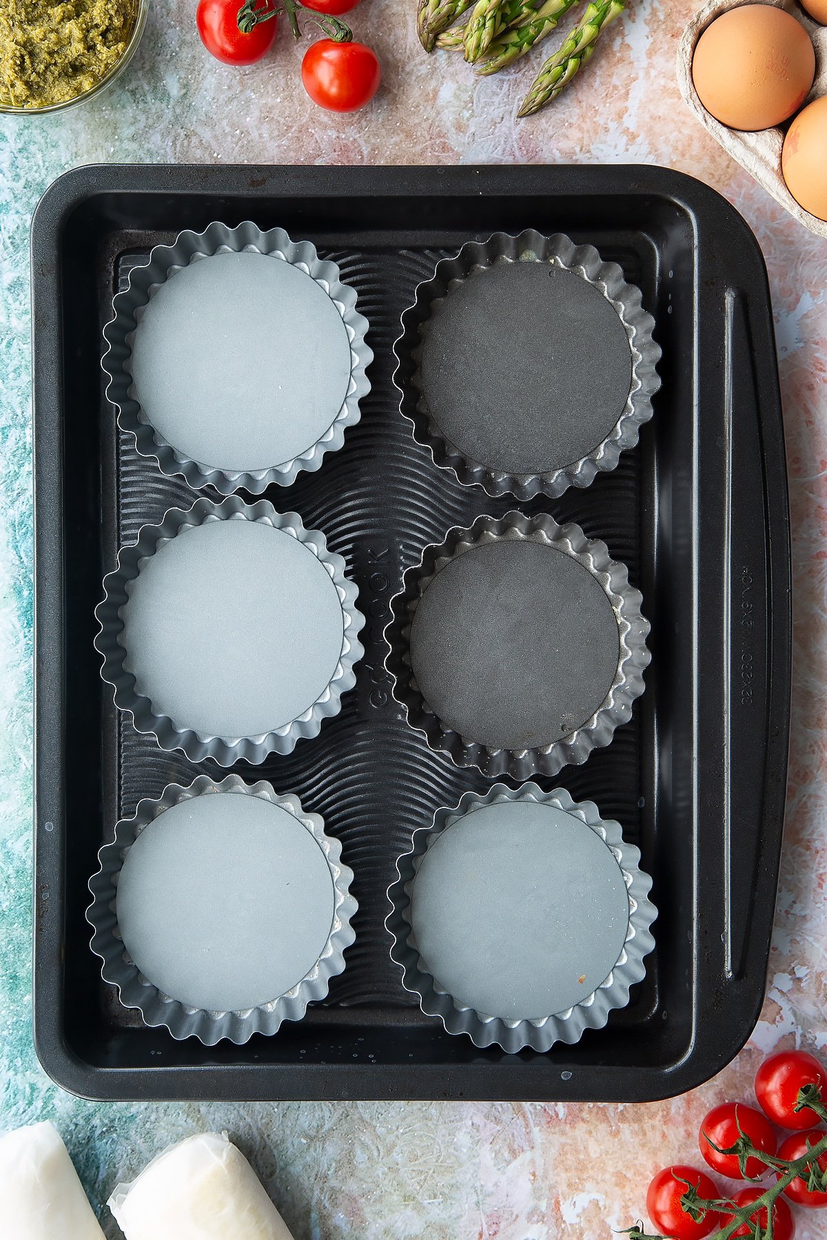 Six tartlet tins on a baking tray. Ingredients to make asparagus tartlets surround the tray.