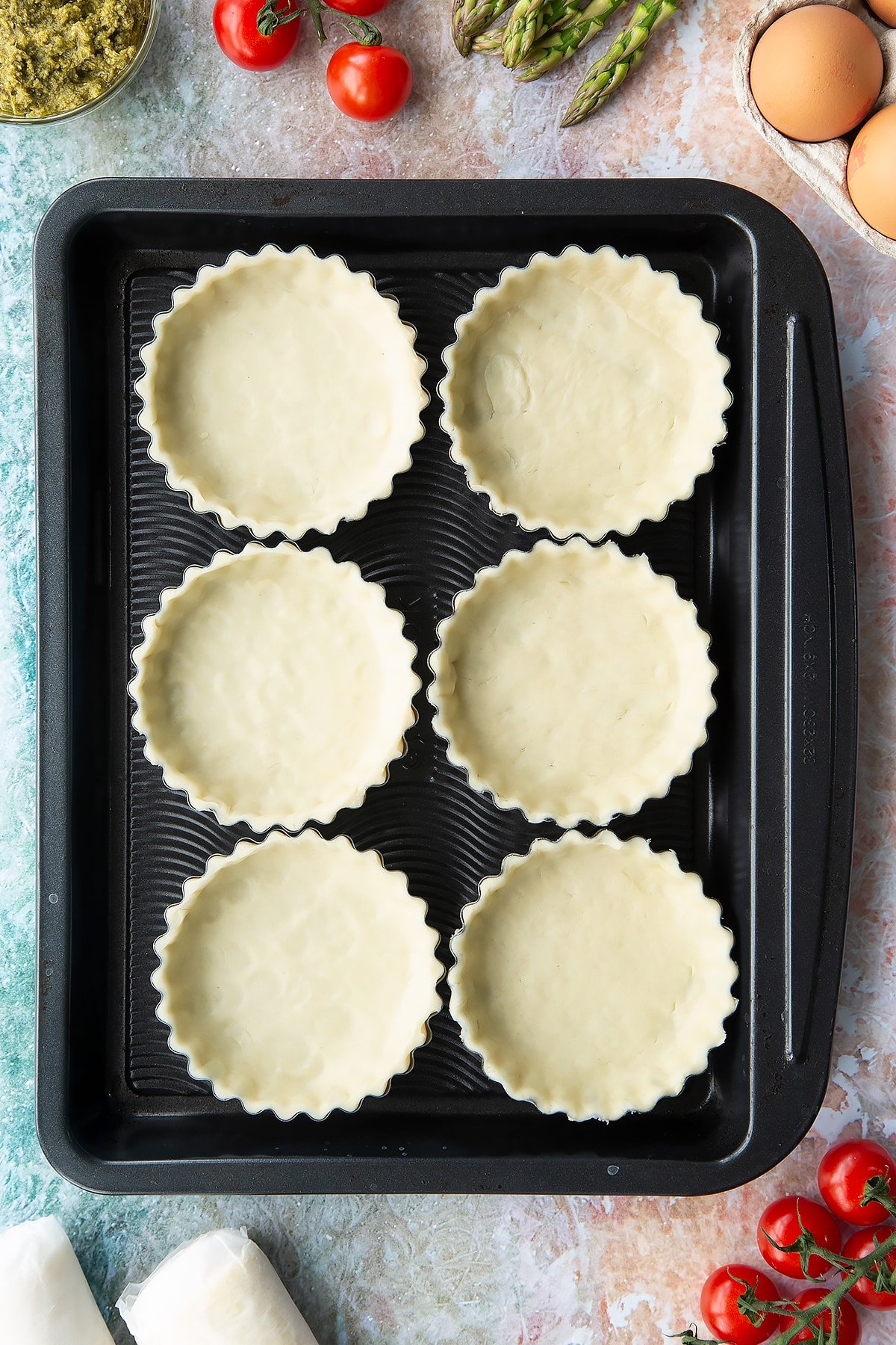 Six tartlet tins on a baking tray. Pastry is fitted and trimmed in the tins. Ingredients to make asparagus tartlets surround the tray.