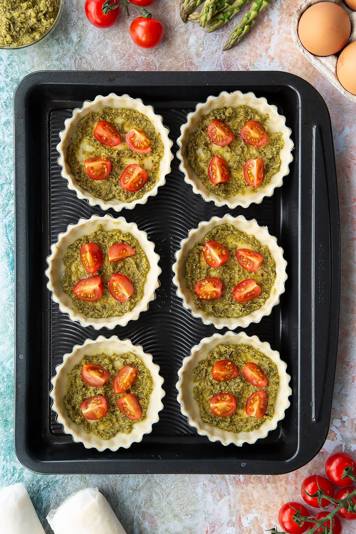 Six tartlet tins on a baking tray. Pastry is fitted and trimmed in the tins. The bottoms are spread with pesto and topped with cherry tomato pieces. Ingredients to make asparagus tartlets surround the tray.