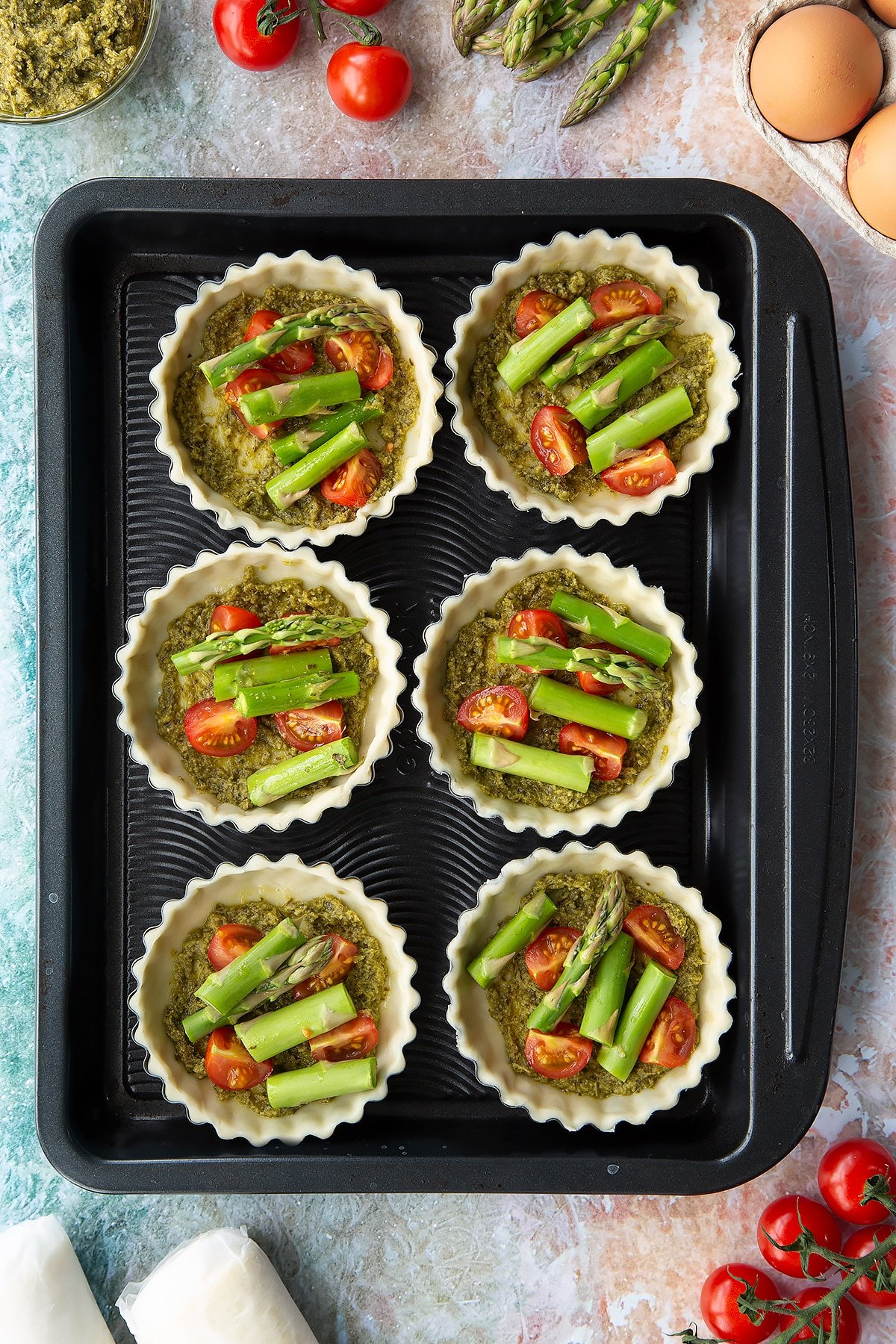 Six tartlet tins on a baking tray. Pastry is fitted and trimmed in the tins. The bottoms are spread with pesto and topped with cherry tomato and asparagus pieces. Ingredients to make asparagus tartlets surround the tray.