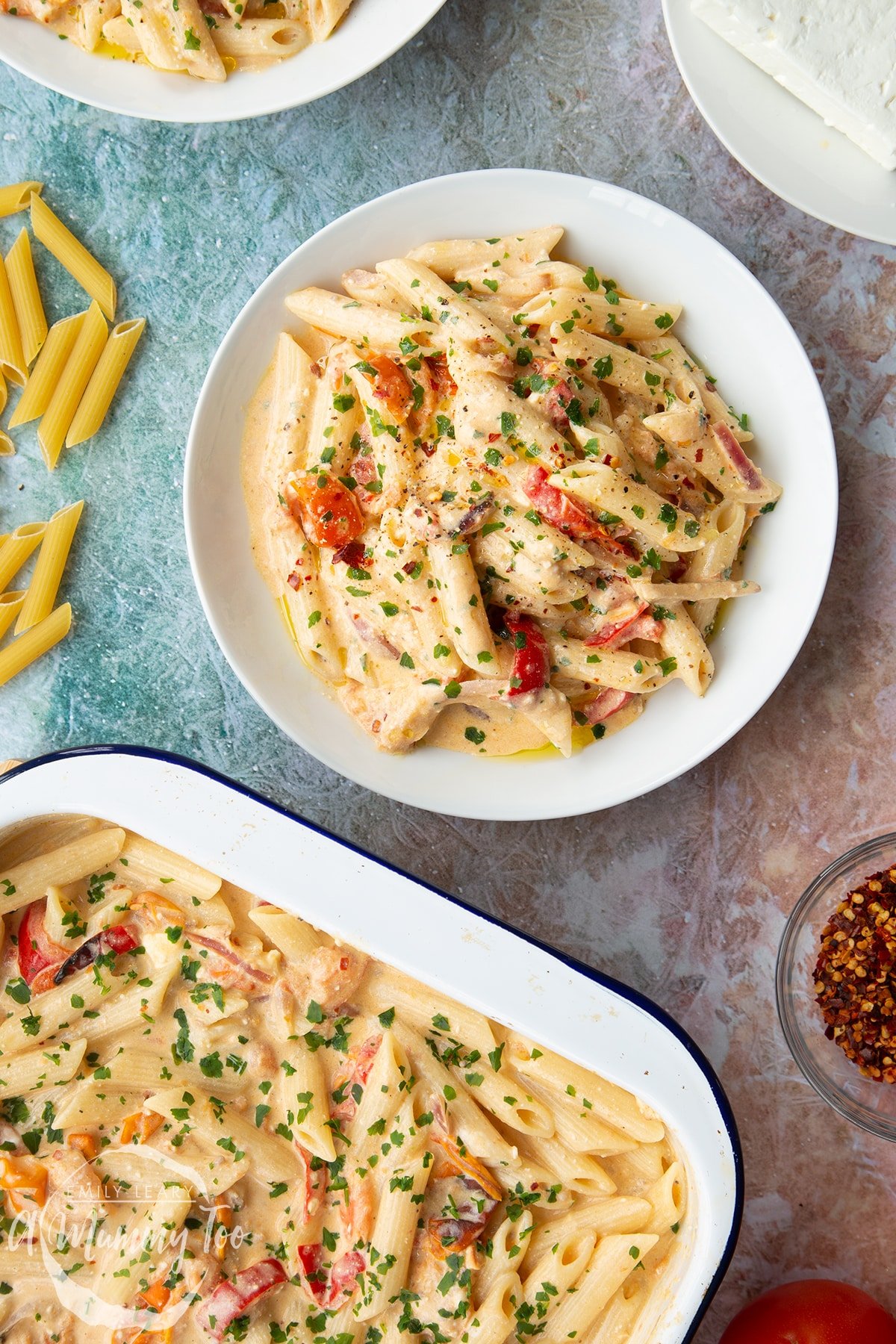 Baked feta pasta in a white bowl, garnished with parsley and chilli flakes. A tray of more pasta is shown to the side.