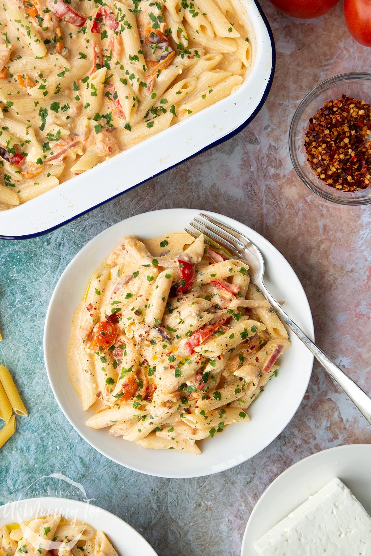 Baked feta pasta in a white bowl, garnished with parsley and chilli flakes. A fork rests in the bowl and a tray of more pasta is shown to the side.