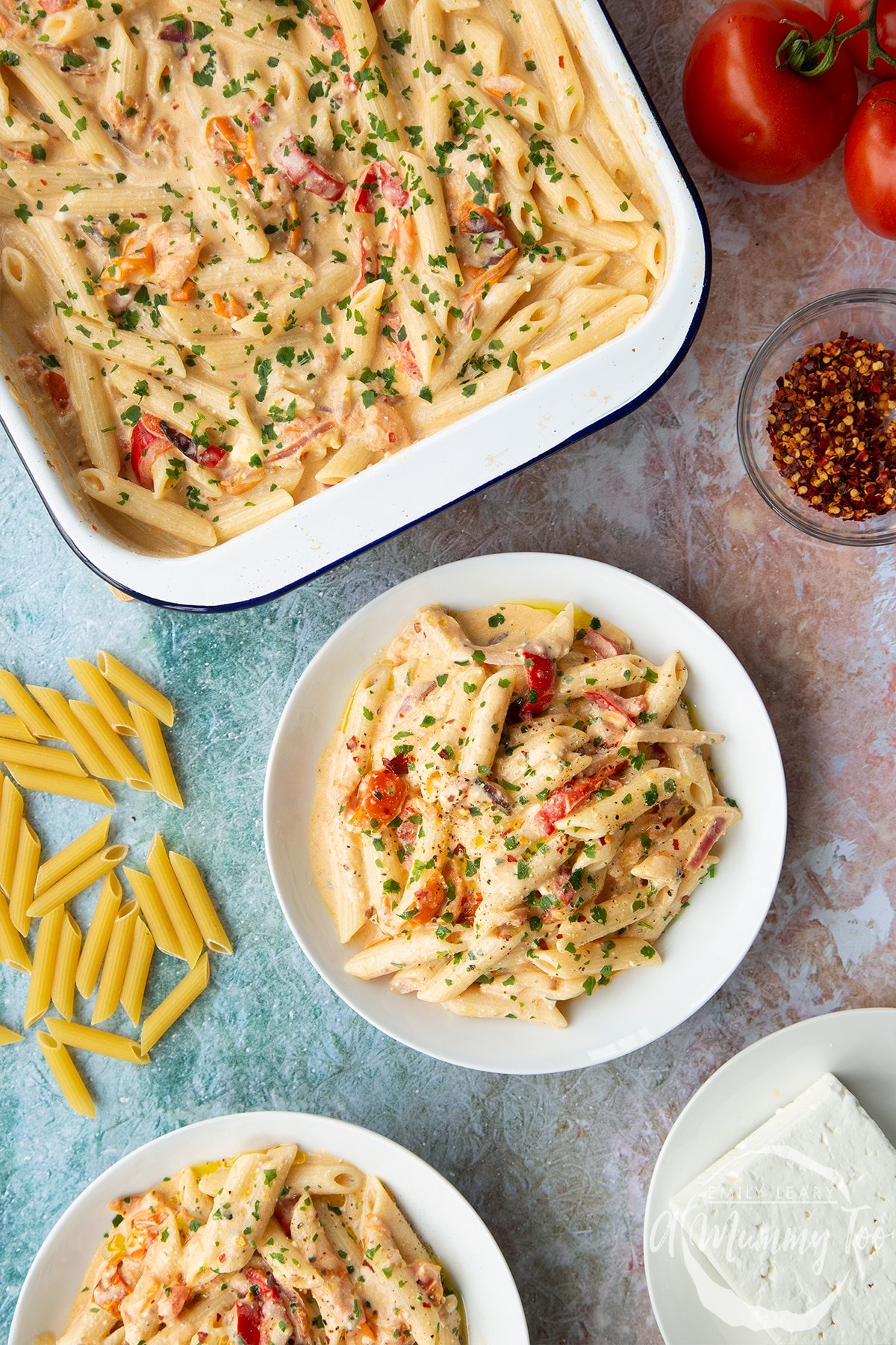 Baked feta pasta in a white bowl, garnished with parsley and chilli flakes. A tray of more pasta is shown beside it.