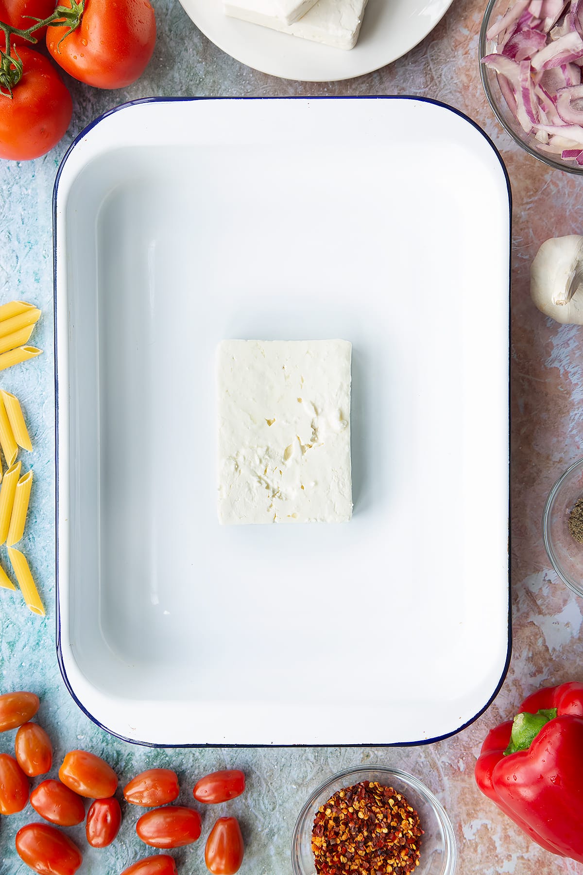 A block of feta in a large white roasting tray. Ingredients to make baked feta pasta surround the tray.