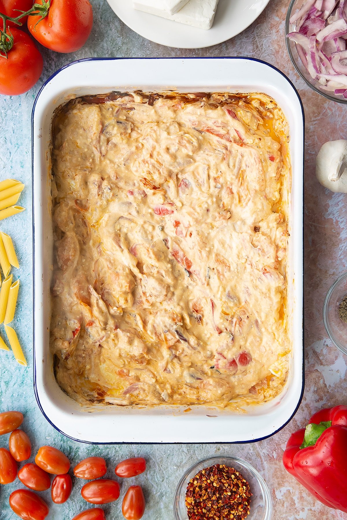 Baked feta, roasted vegetables and cream cheese stirred together in a large white roasting tray. Ingredients to make baked feta pasta surround the tray.