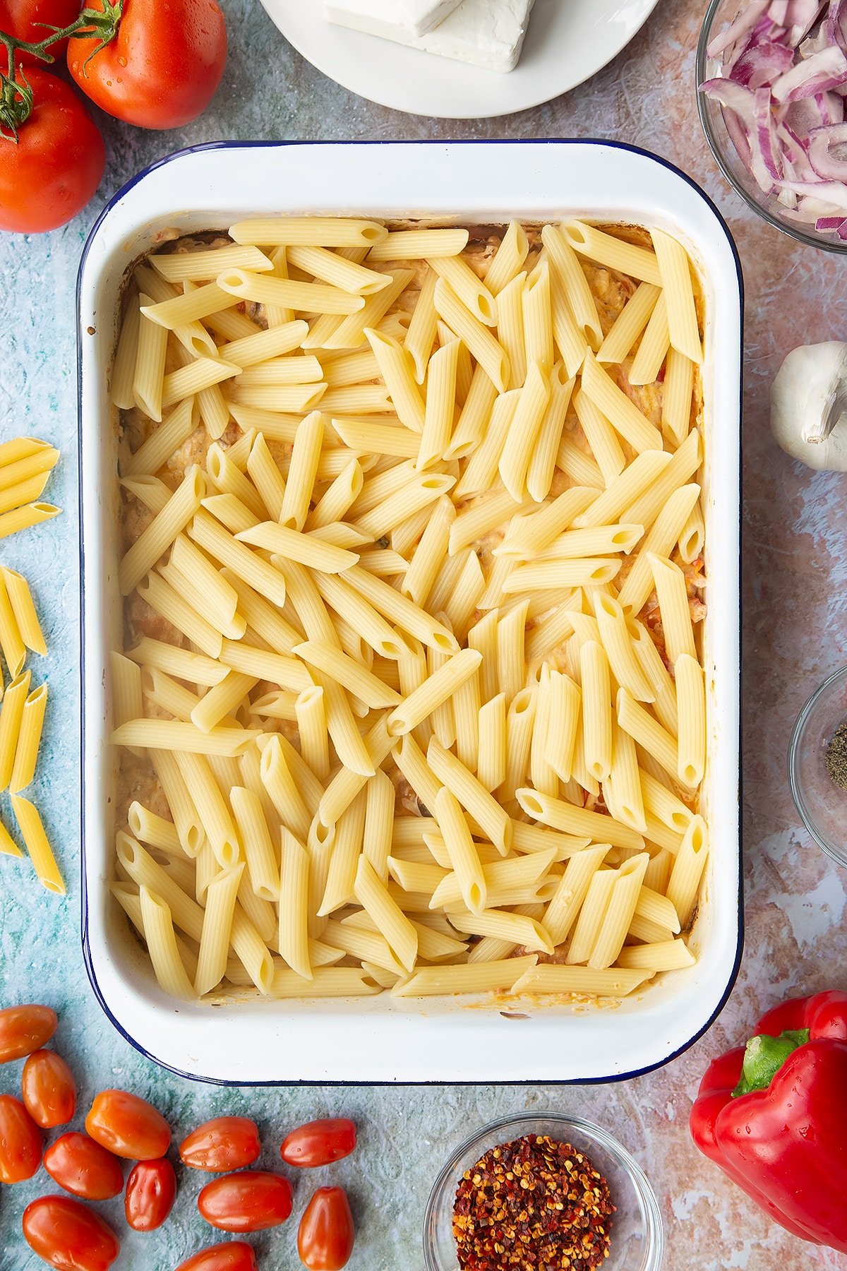 Baked feta, roasted vegetables and cream cheese with penne on top in a large white roasting tray. Ingredients to make baked feta pasta surround the tray.