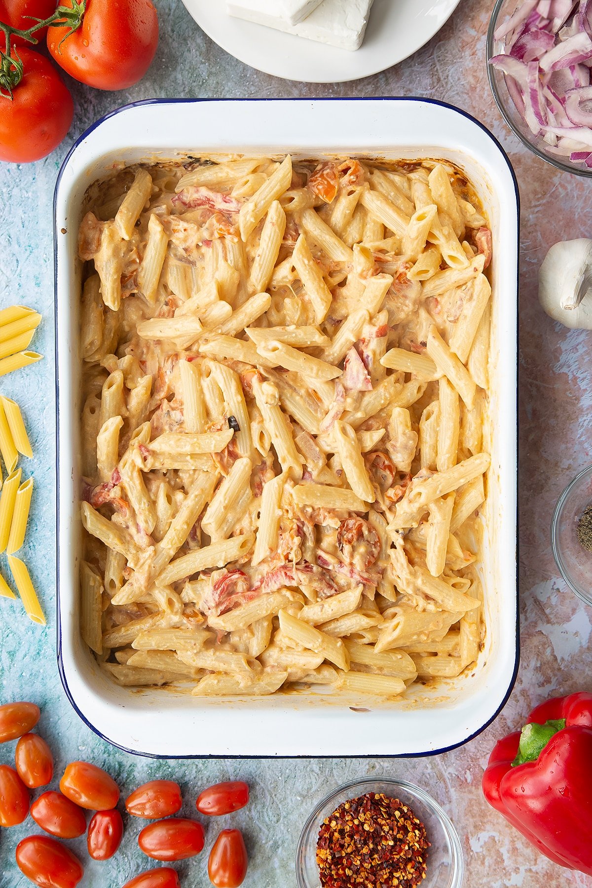 Baked feta, roasted vegetables, cream cheese and penne stirred together in a large white roasting tray. Ingredients to make baked feta pasta surround the tray.