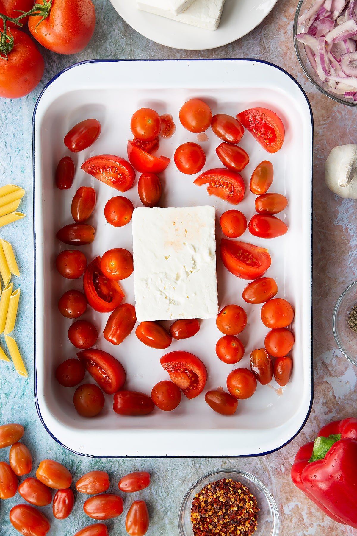 A block of feta surrounded by tomatoes in a large white roasting tray. Ingredients to make baked feta pasta surround the tray.