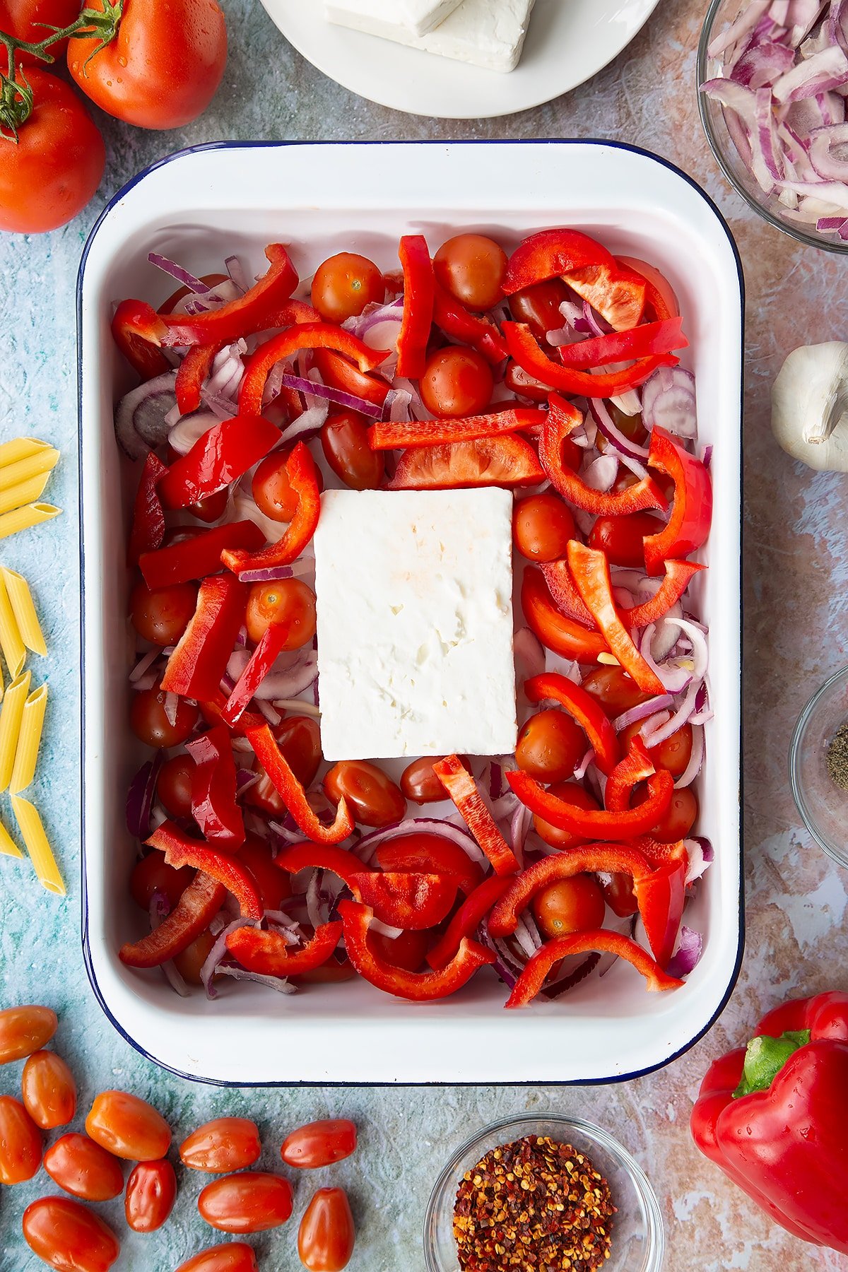 A block of feta surrounded by tomatoes, red pepper and red onion slices in a large white roasting tray. Ingredients to make baked feta pasta surround the tray.