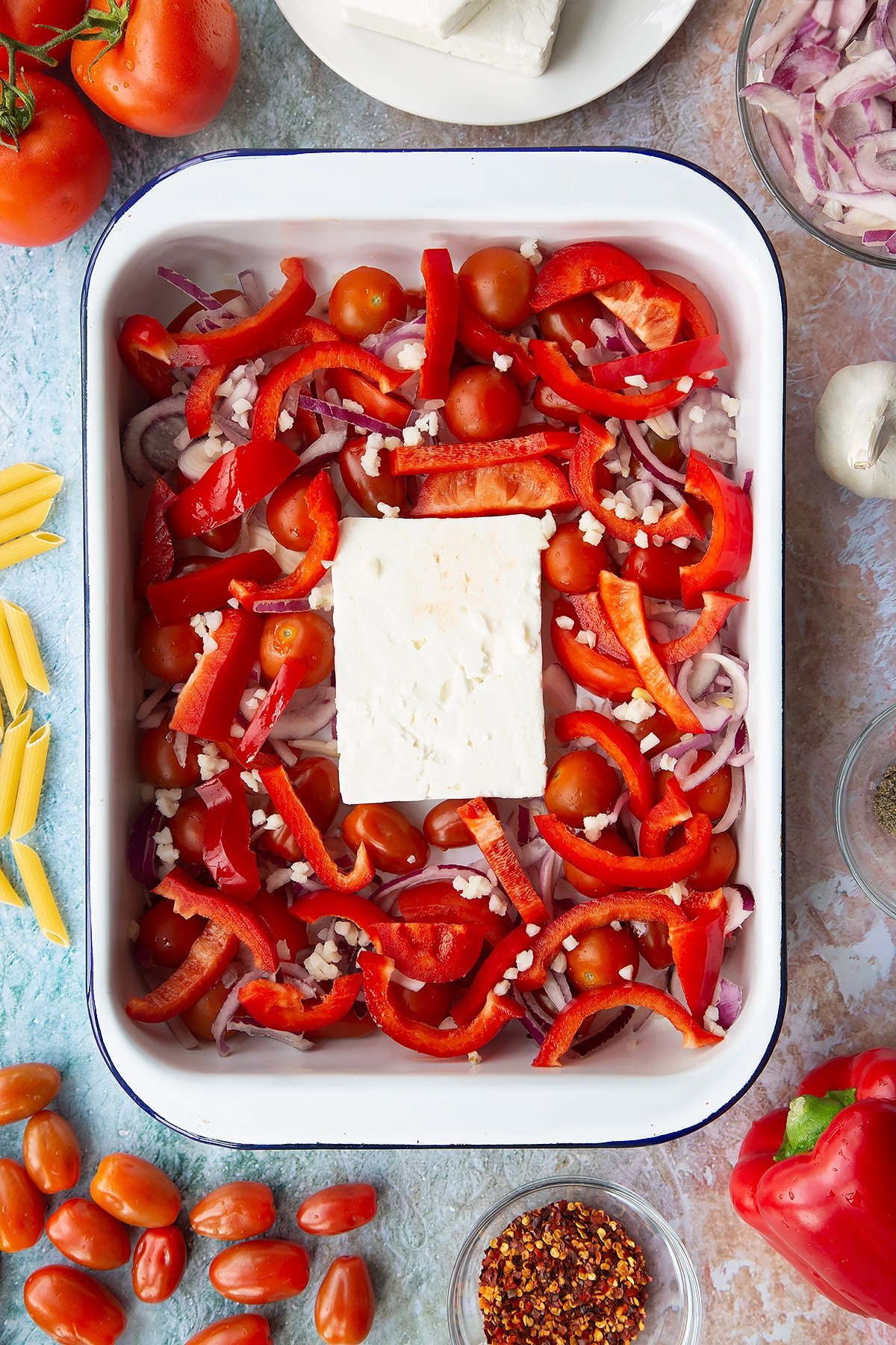 A block of feta surrounded by tomatoes, red pepper, red onion and garlic in a large white roasting tray. Ingredients to make baked feta pasta surround the tray.