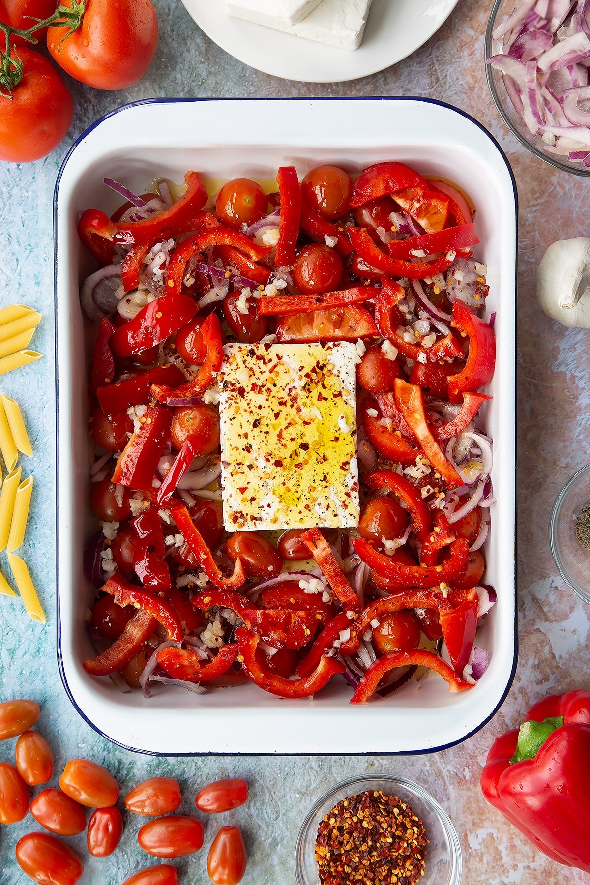 A block of feta surrounded by tomatoes, red pepper, red onion and garlic in a large white roasting tray, topped with olive oil, chilli flakes, salt and pepper. Ingredients to make baked feta pasta surround the tray.