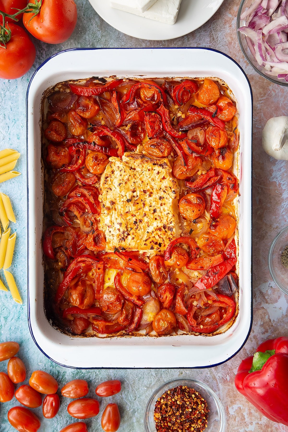 A block of baked feta surrounded by roasted tomatoes, red pepper, red onion and garlic in a large white roasting tray. Ingredients to make baked feta pasta surround the tray.