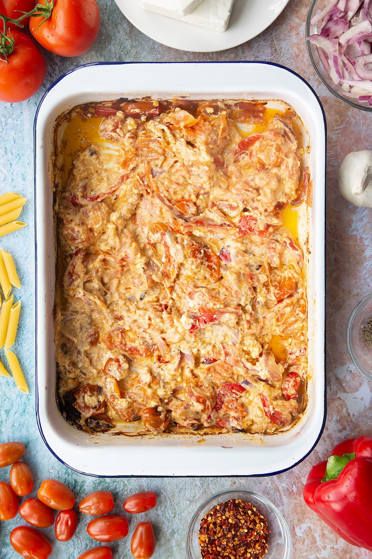 Baked feta and roasted vegetables stirred together in a large white roasting tray. Ingredients to make baked feta pasta surround the tray.