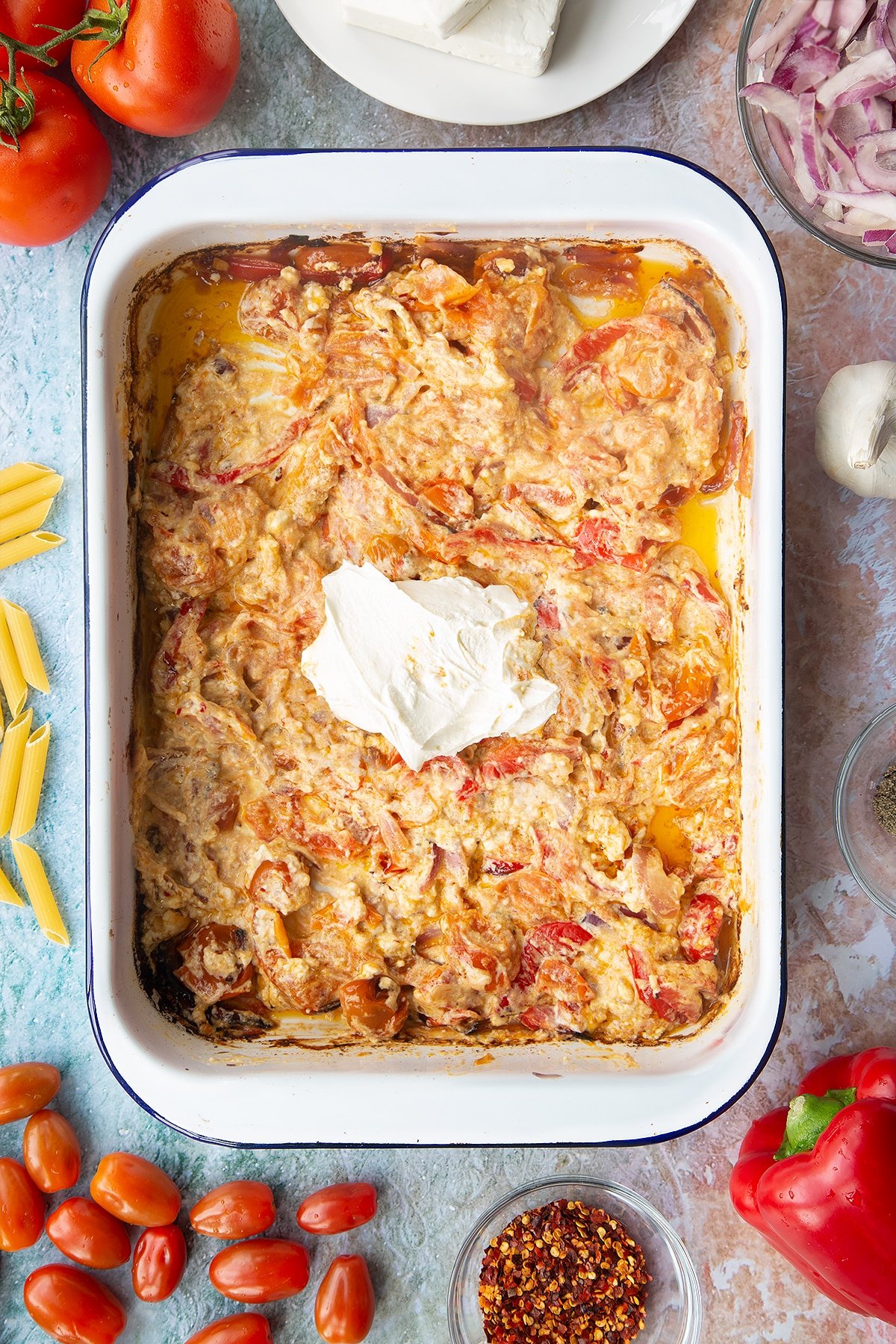 Baked feta and roasted vegetables stirred together in a large white roasting tray with a dollop of cream cheese on top. Ingredients to make baked feta pasta surround the tray.