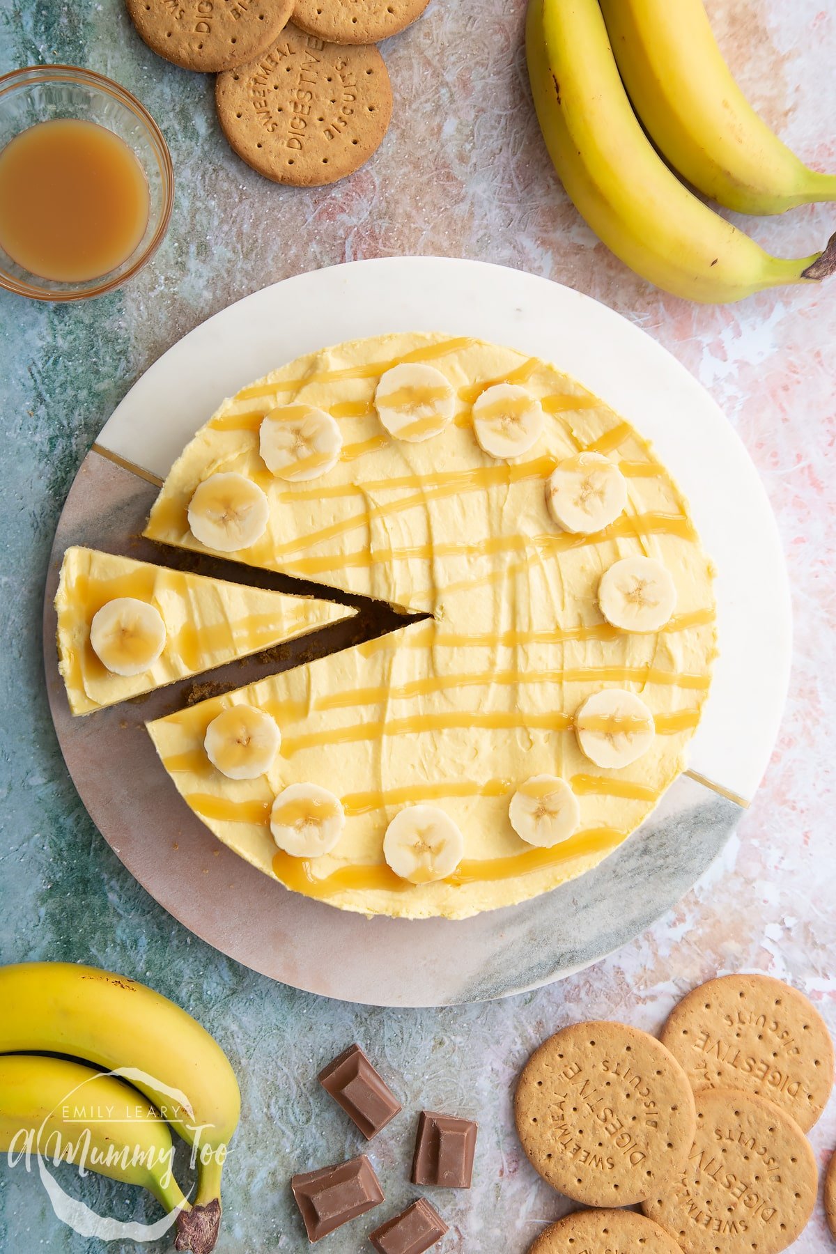 Banoffee pie cheesecake topped with banana slices and drizzled with toffee sauce on a white and pink marble board. A slice is partly removed.