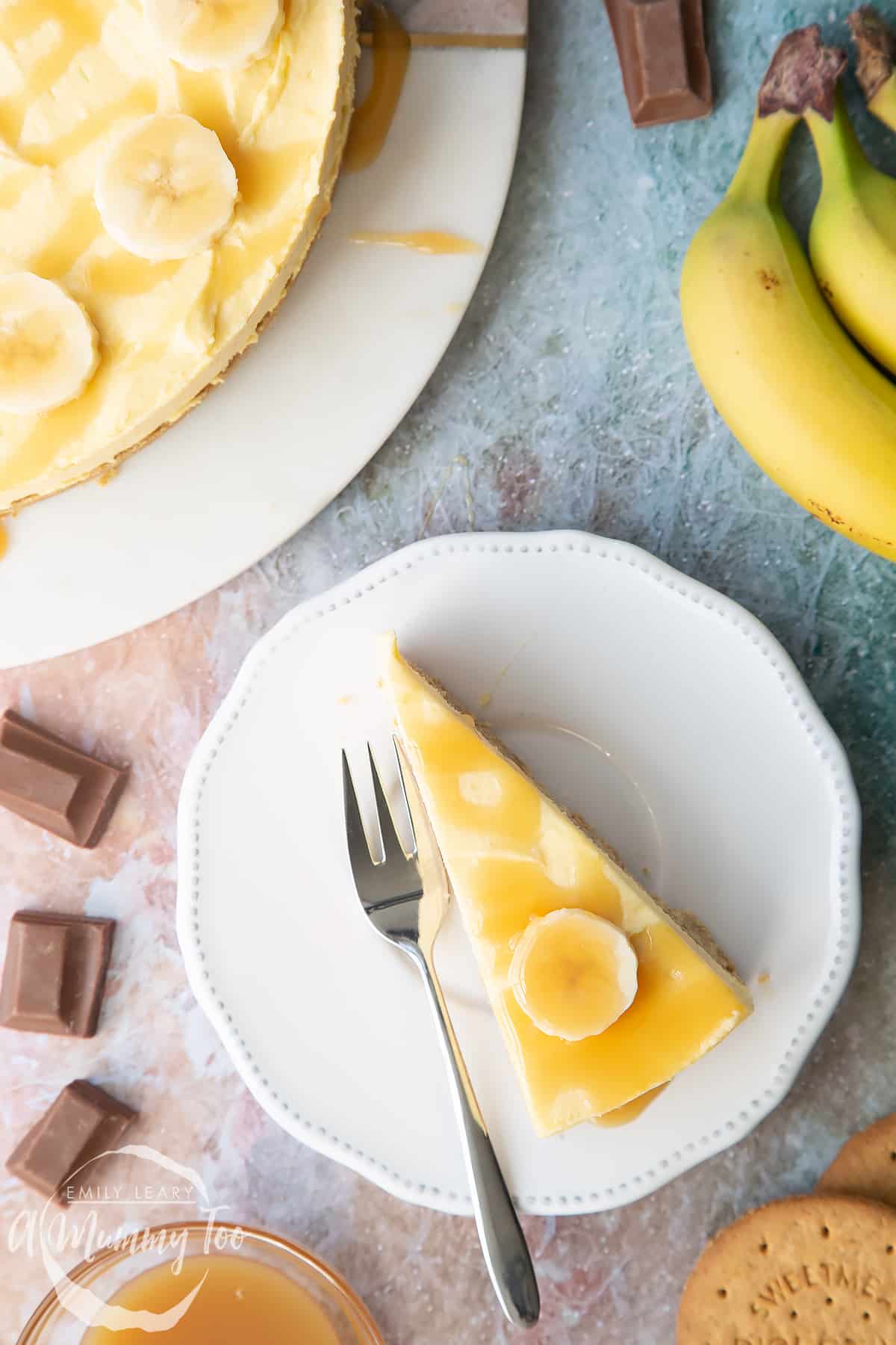 A slice of banoffee pie cheesecake on a white plate with a fork resting beside it, shown from above.