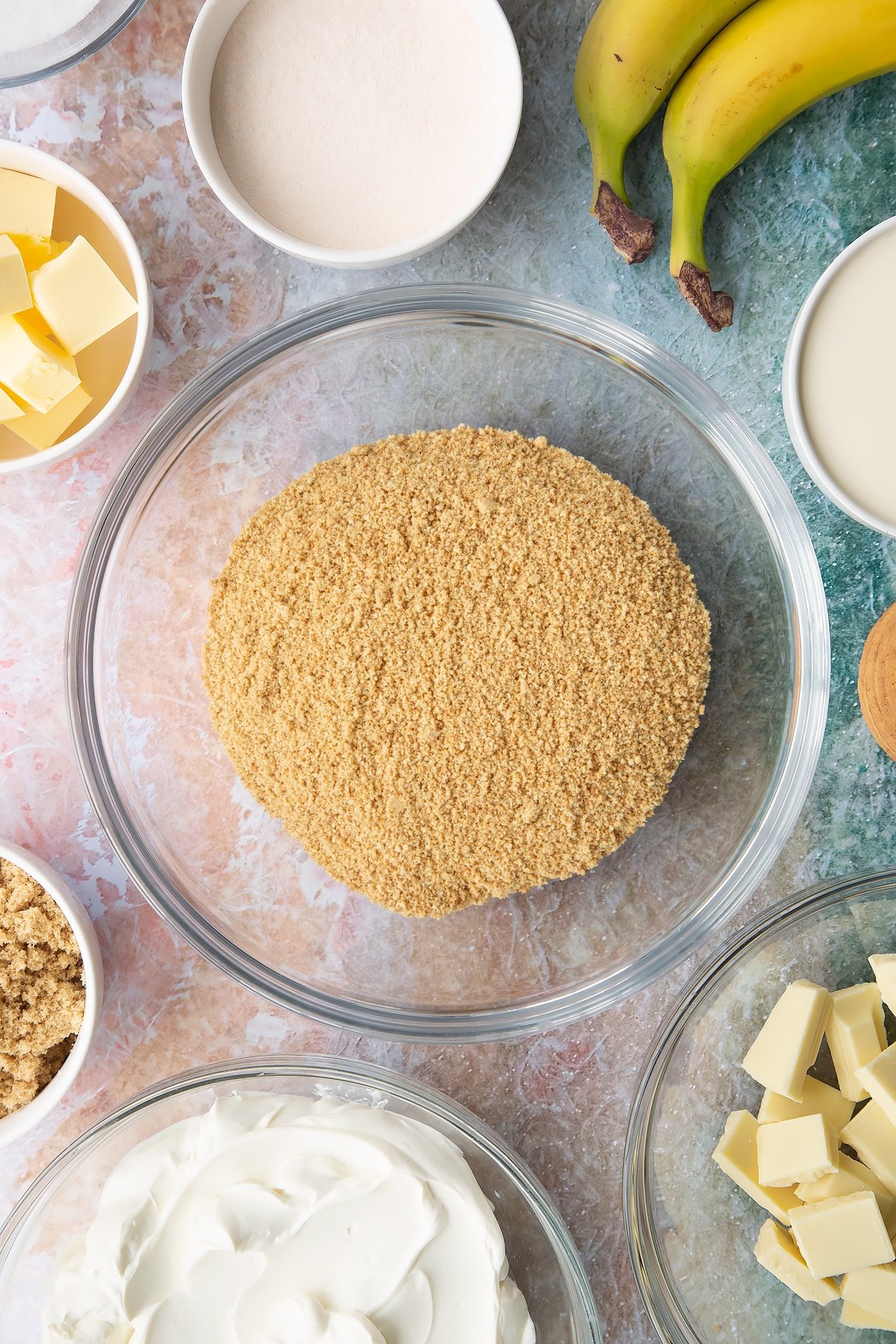 Crumbled biscuits in a bowl. Ingredients to make banoffee pie cheesecake surround the bowl.