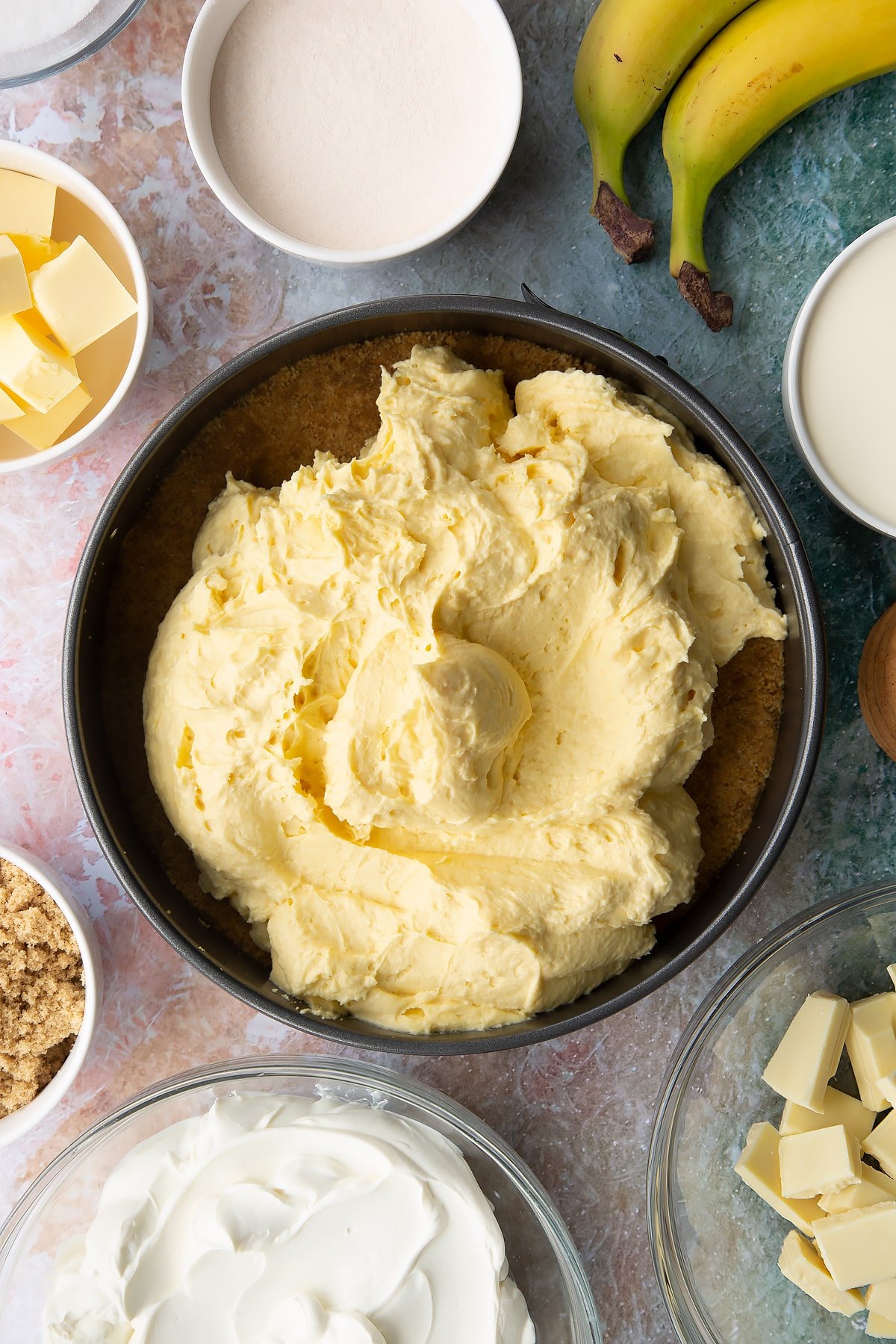 Banoffee cheesecake mix tipped into a tin on top of the biscuit base. Ingredients to make banoffee pie cheesecake surround the tin.