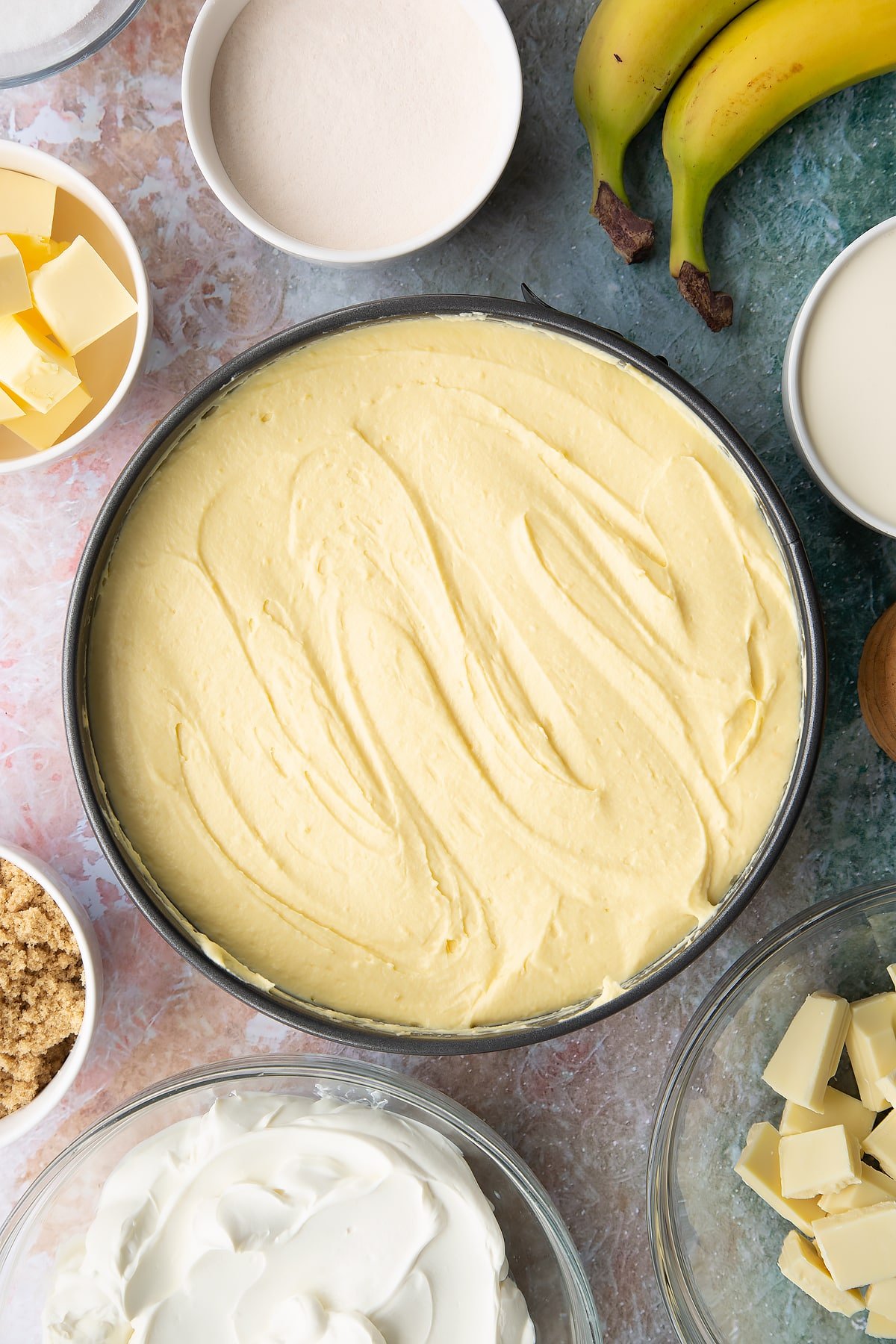 Banoffee cheesecake mix spread on top of the biscuit base. Ingredients to make banoffee pie cheesecake surround the tin.