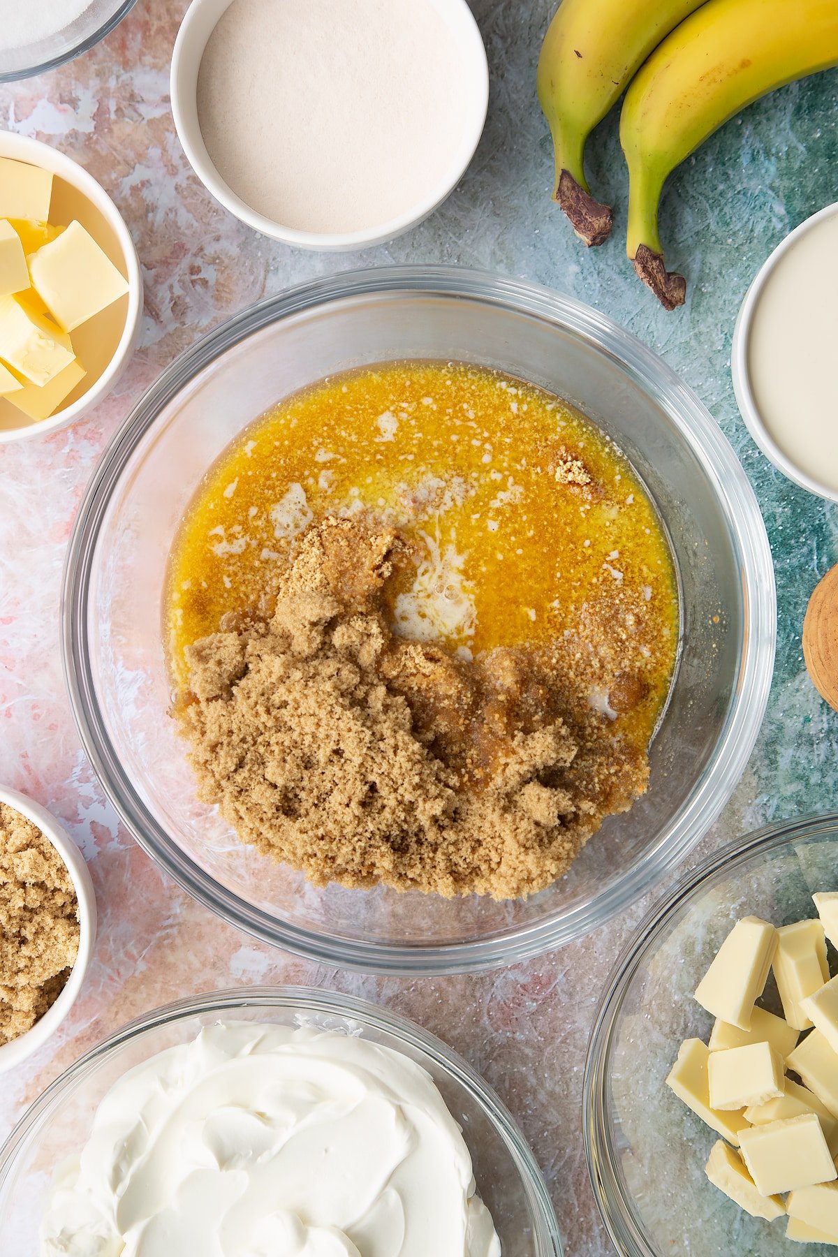 Crumbled biscuits and melted butter in a bowl. Ingredients to make banoffee pie cheesecake surround the bowl.