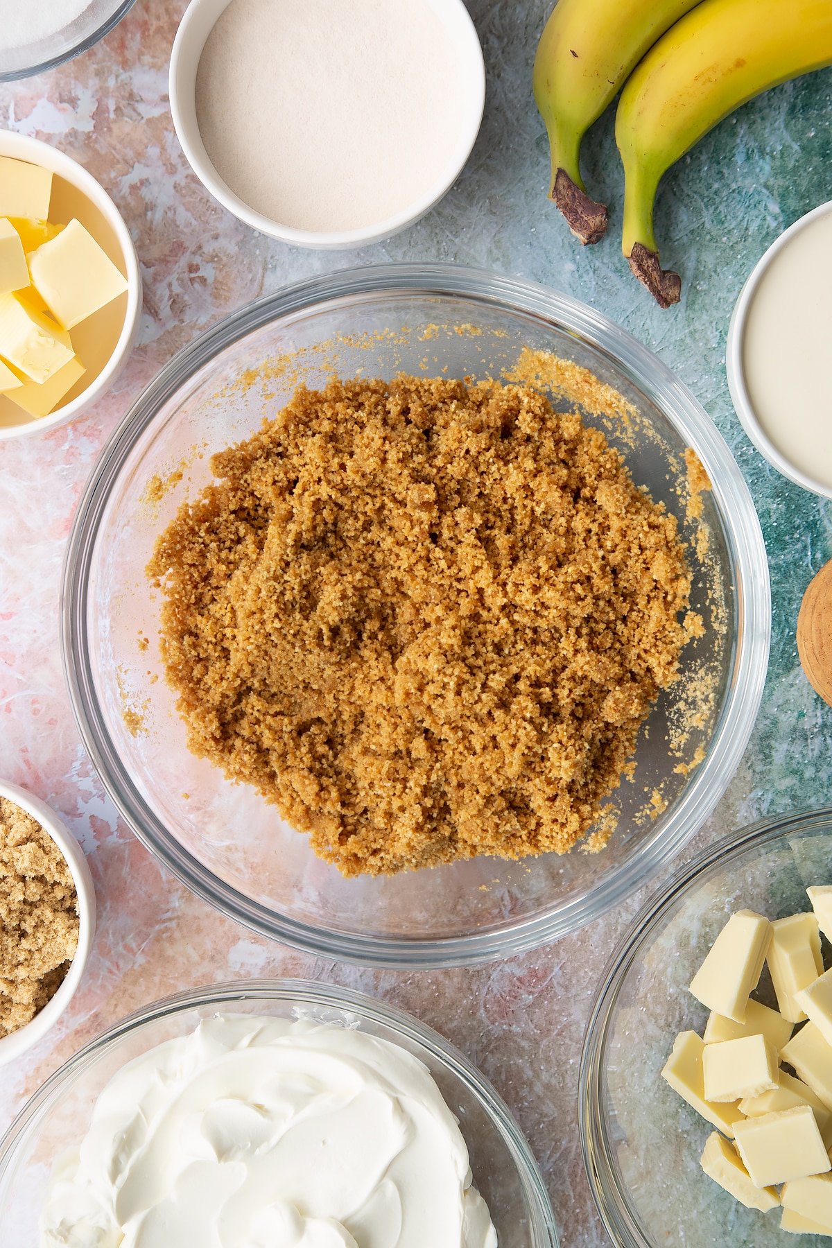 Crumbled biscuits and melted butter mixed together in a bowl. Ingredients to make banoffee pie cheesecake surround the bowl.