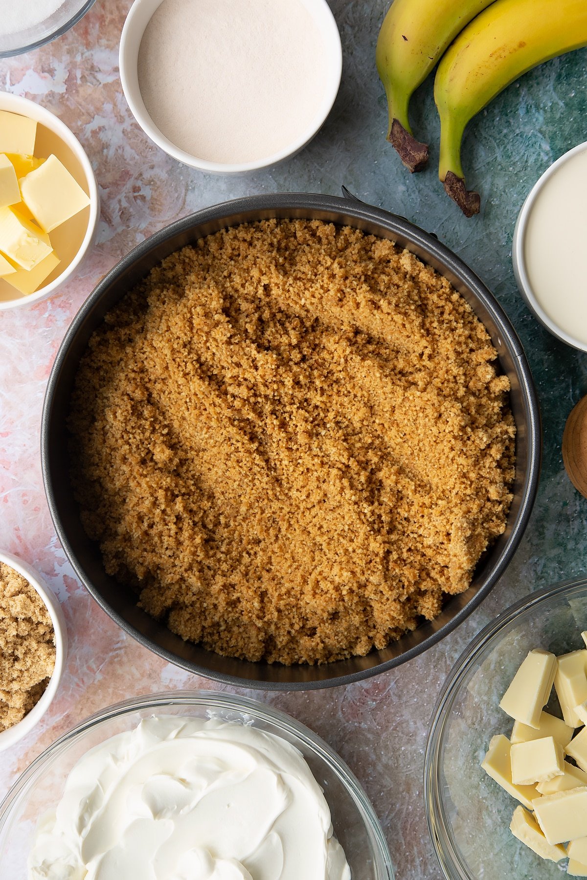 Crumbled biscuits and melted butter mixed together and tipped into a large cake tin. Ingredients to make banoffee pie cheesecake surround the tin.