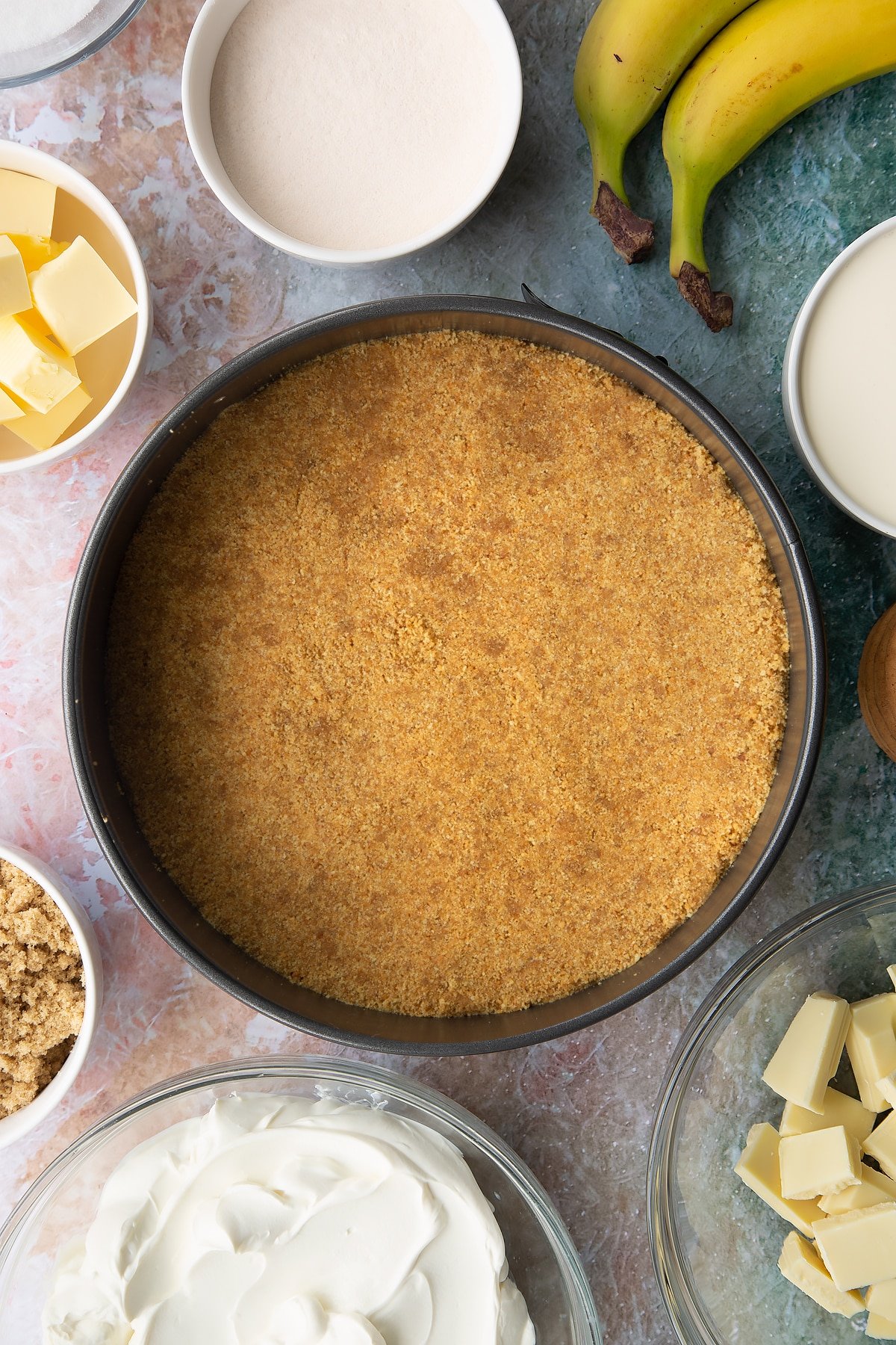 Crumbled biscuits and melted butter mixed together and pressed into the base of a large cake tin. Ingredients to make banoffee pie cheesecake surround the tin.