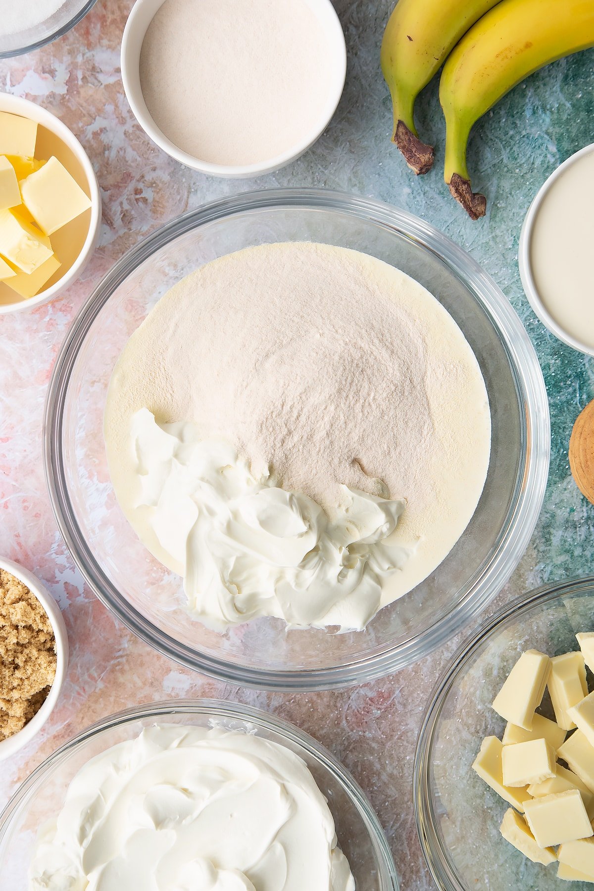 Cream, cream cheese, sugar and banana pudding powder in a bowl. Ingredients to make banoffee pie cheesecake surround the bowl.