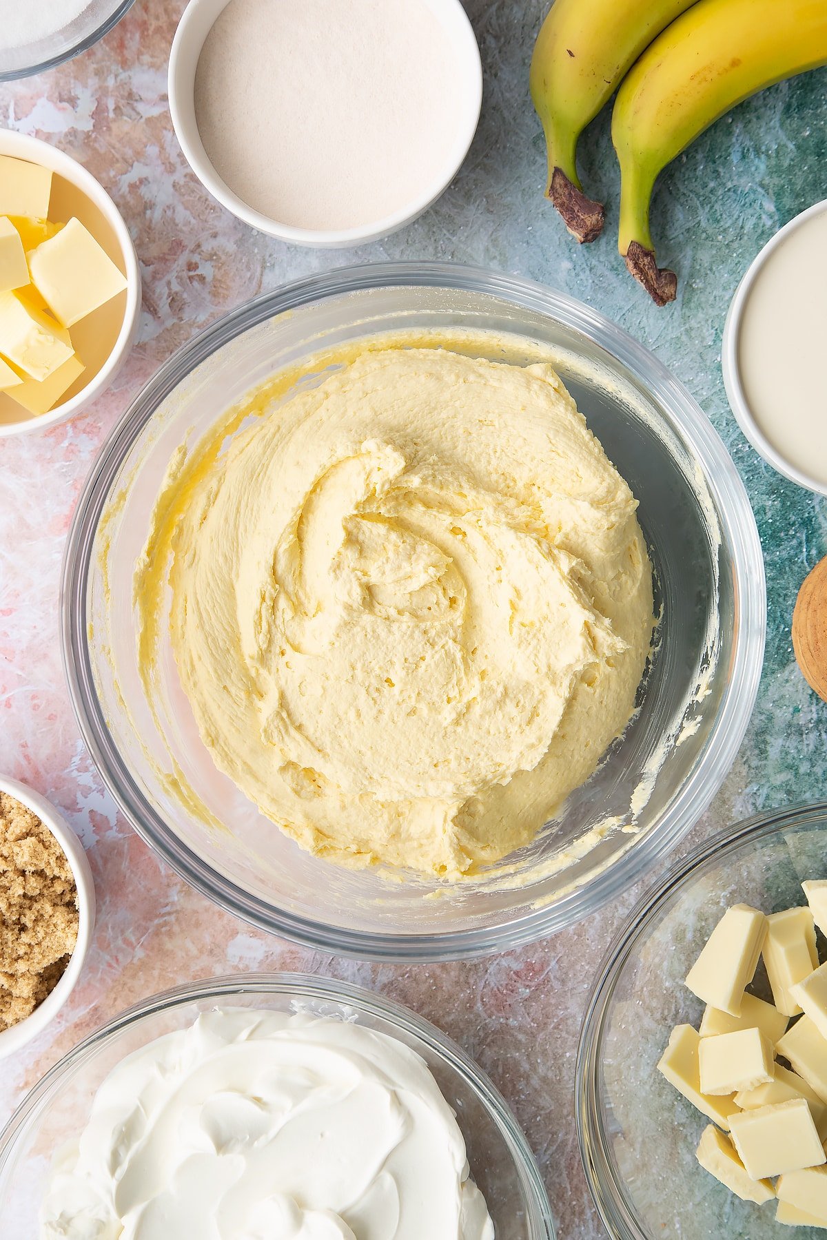 Cream, cream cheese, sugar and banana pudding powder whisked together in a bowl. Ingredients to make banoffee pie cheesecake surround the bowl.