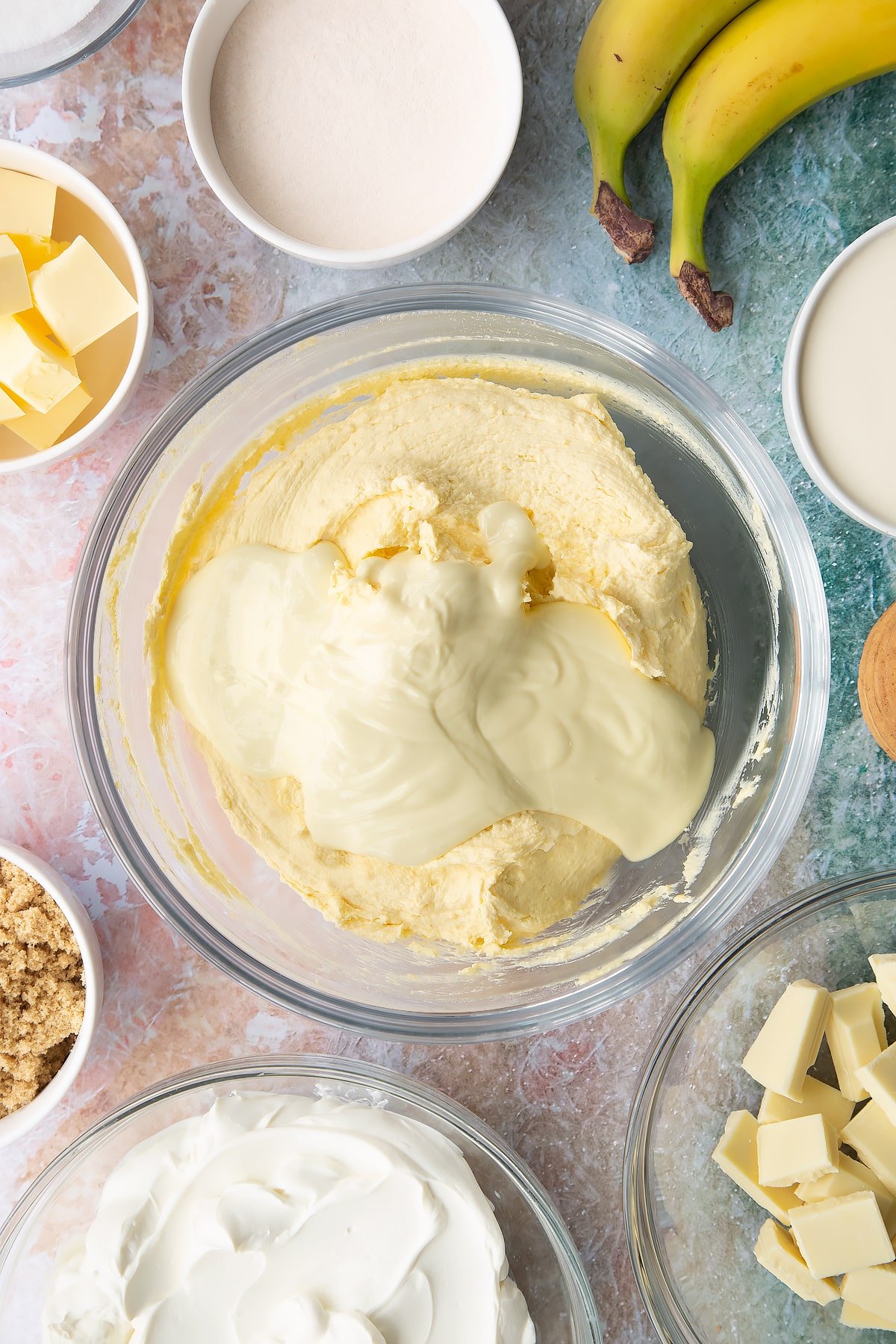 Cream, cream cheese, sugar and banana pudding powder whisked together in a bowl with melted white chocolate on top. Ingredients to make banoffee pie cheesecake surround the bowl.